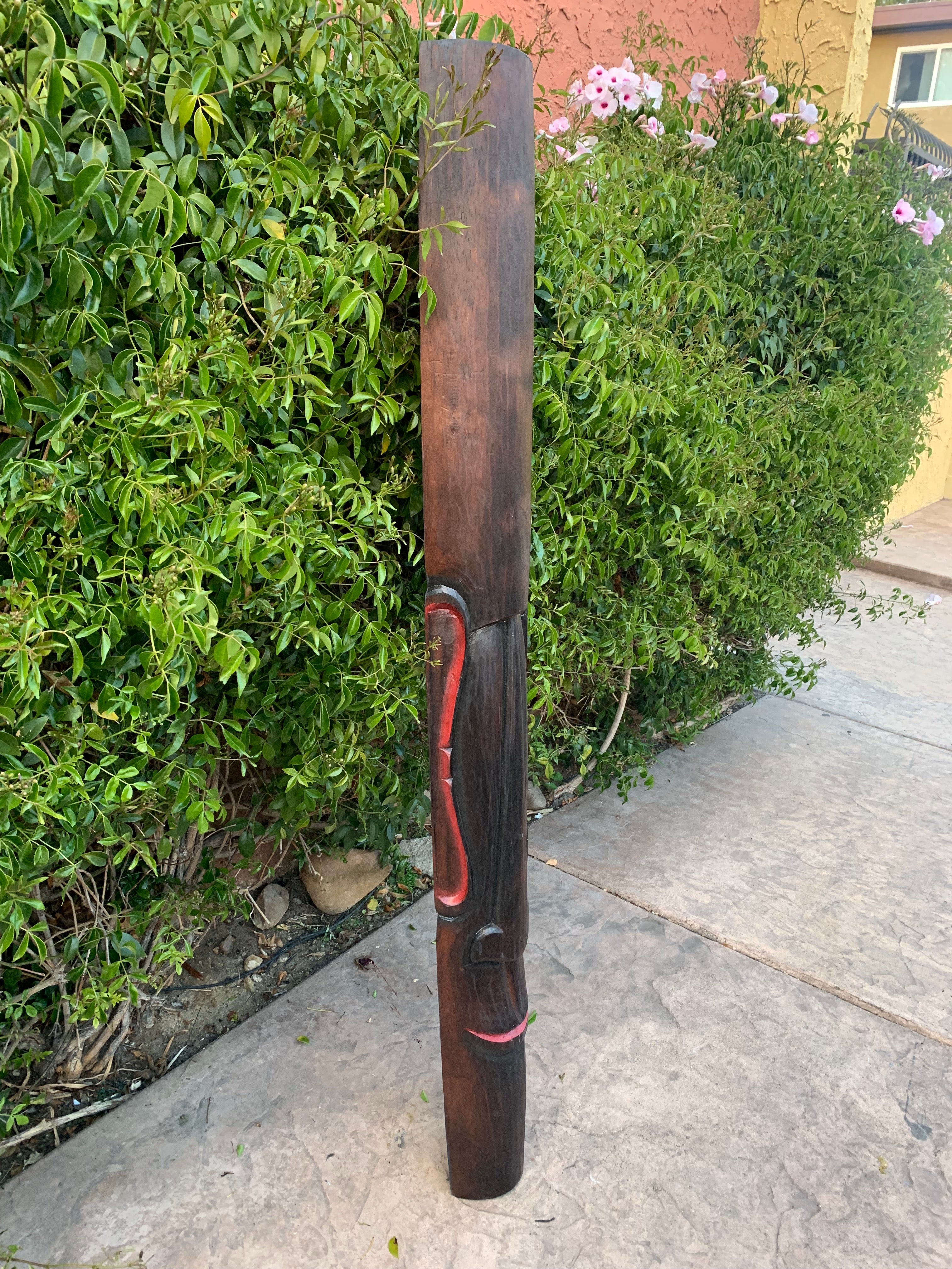 Tall carved wooden post with a facial carving and red accents beside a green hedge and pink flowers