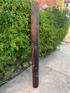 Tall carved wooden post with a facial carving and red accents beside a green hedge and pink flowers