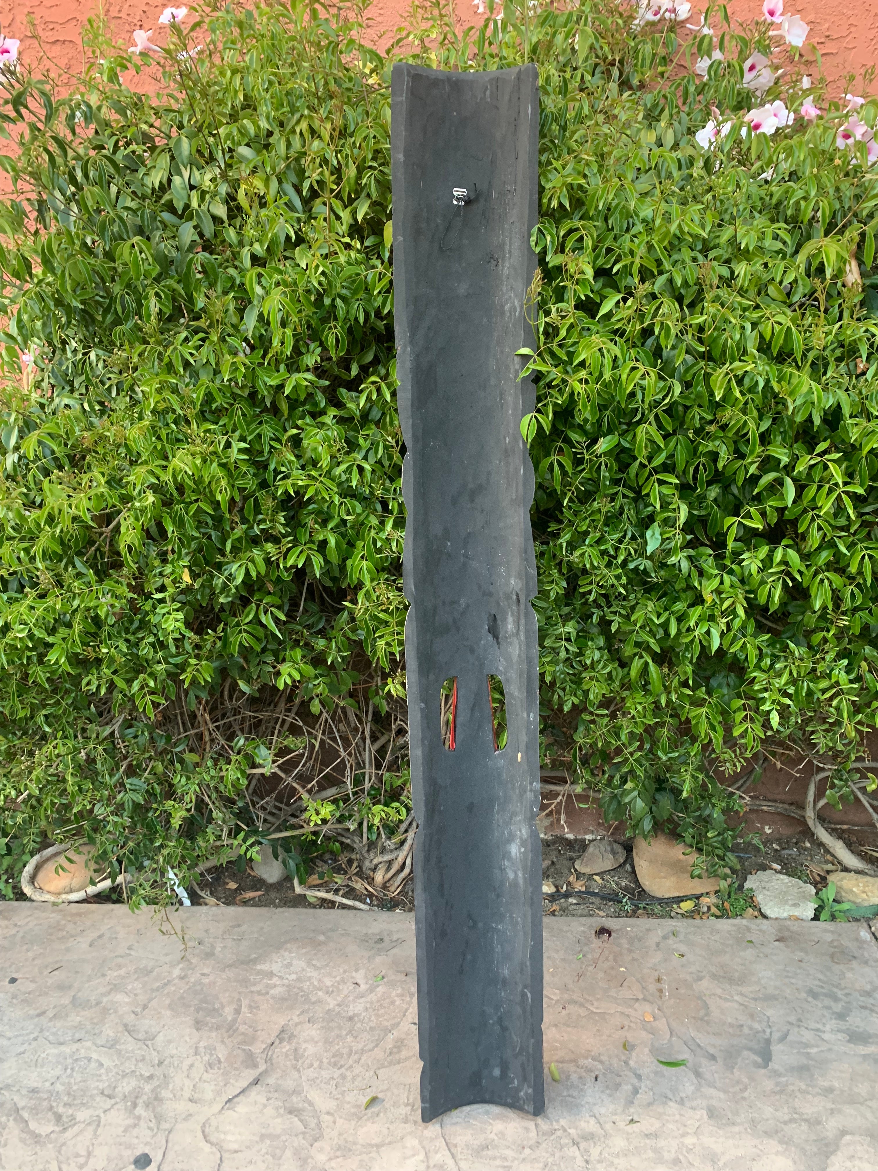 Tall dark slate outdoor sculpture with a small faucet near the top, placed among lush green shrubs against a peach wall