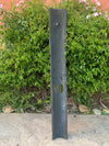 Tall dark slate outdoor sculpture with a small faucet near the top, placed among lush green shrubs against a peach wall