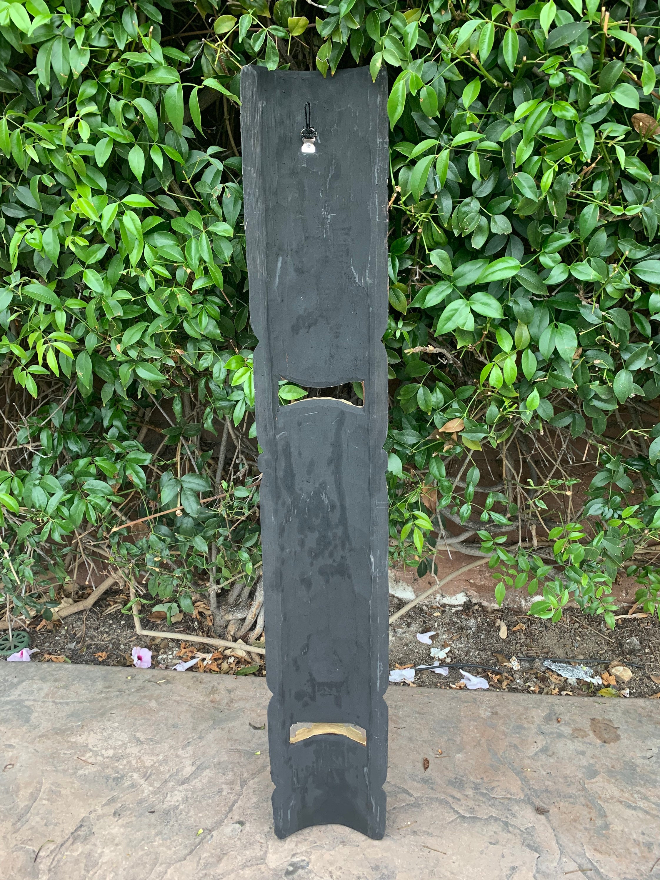 Tall dark slate panel standing upright in a garden with a green hedge, featuring rectangular and curved cutouts.