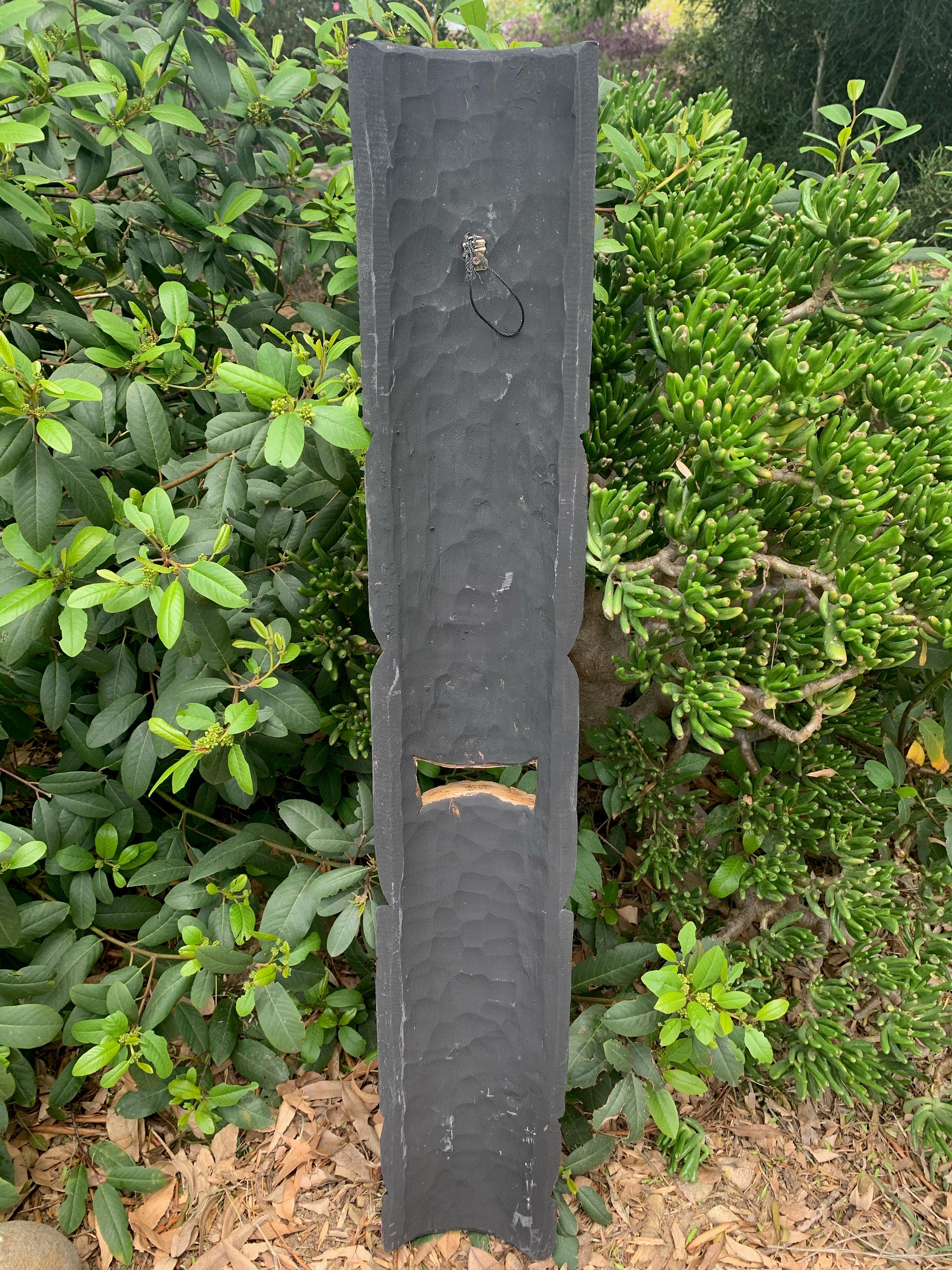 Tall dark slate pillar with a rectangular notch, set among green shrubs in a garden