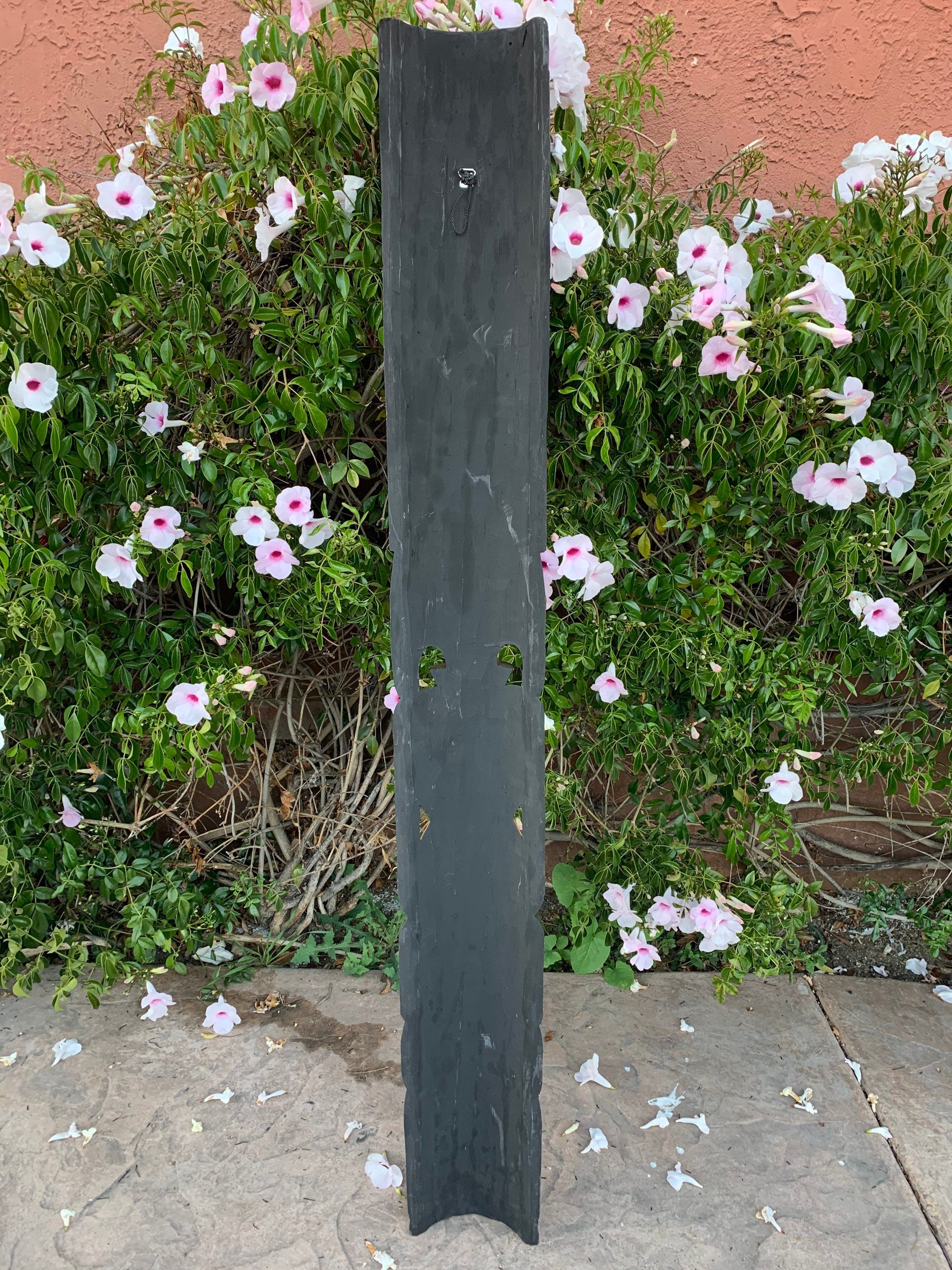 Tall dark weathered wooden post with cut-out holes in a garden with pink and white flowers