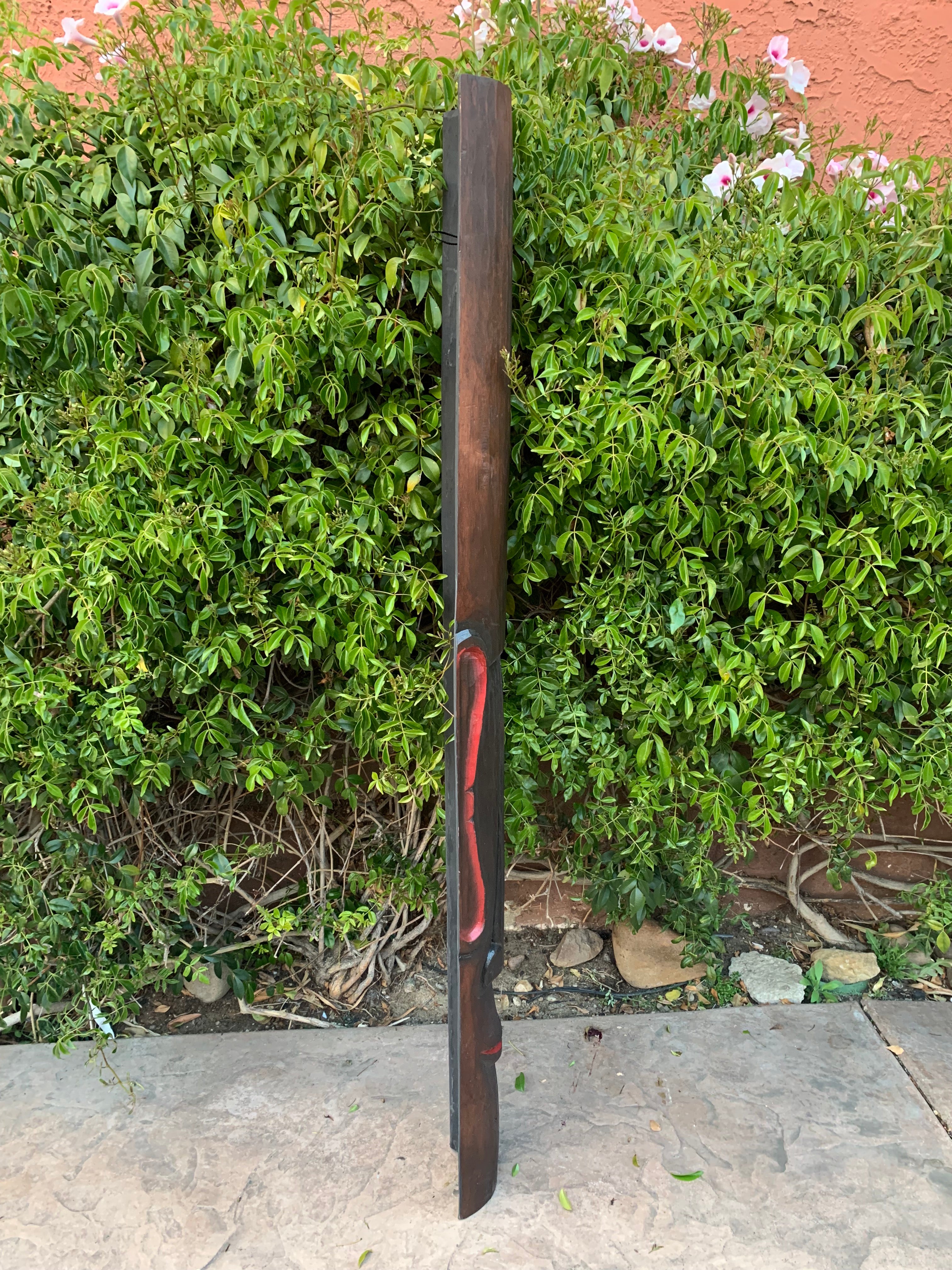 Tall dark wooden post with red carved design leaning against a green hedge.