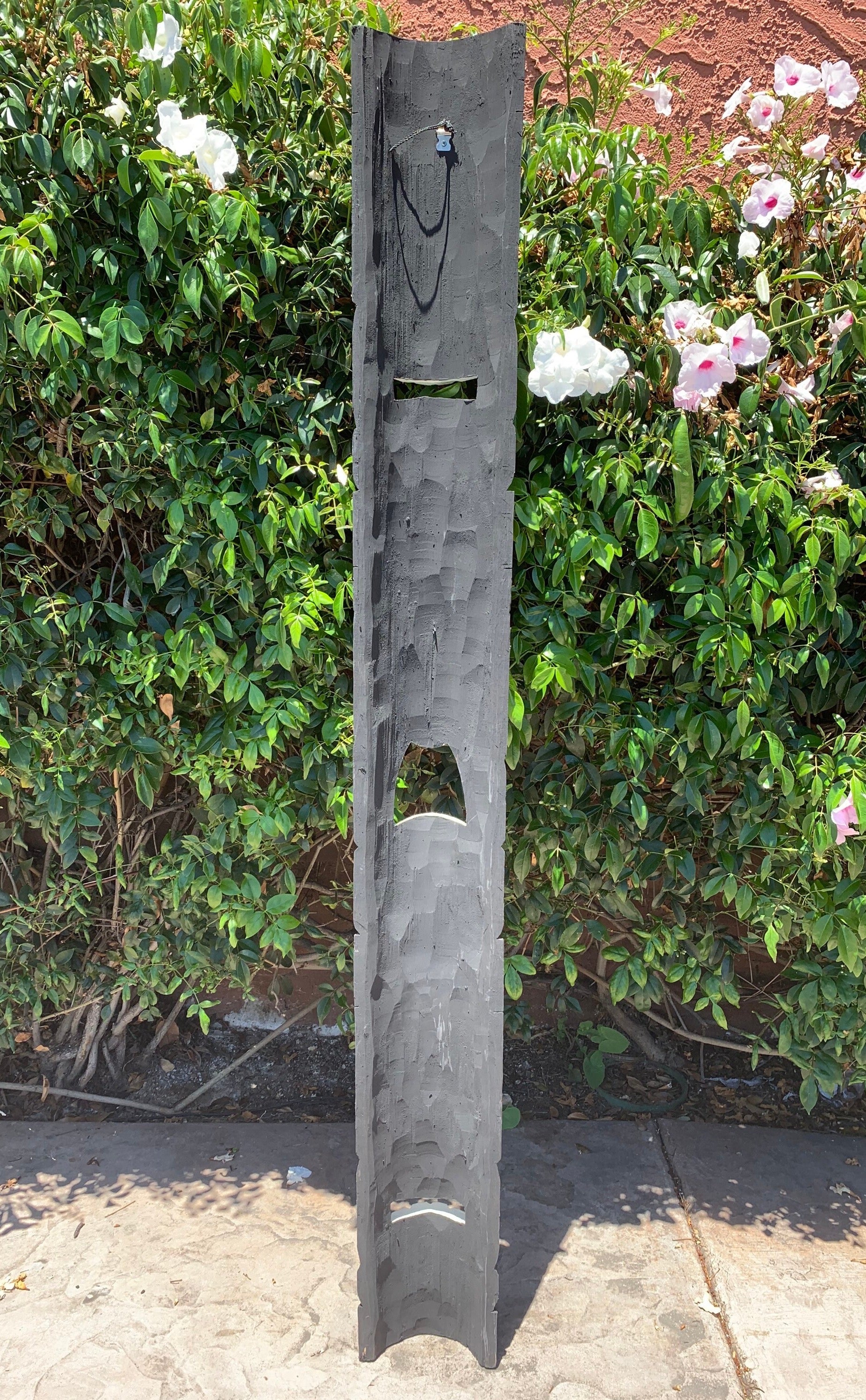 Tall gray wooden sculpture post with geometric cutouts in a garden setting