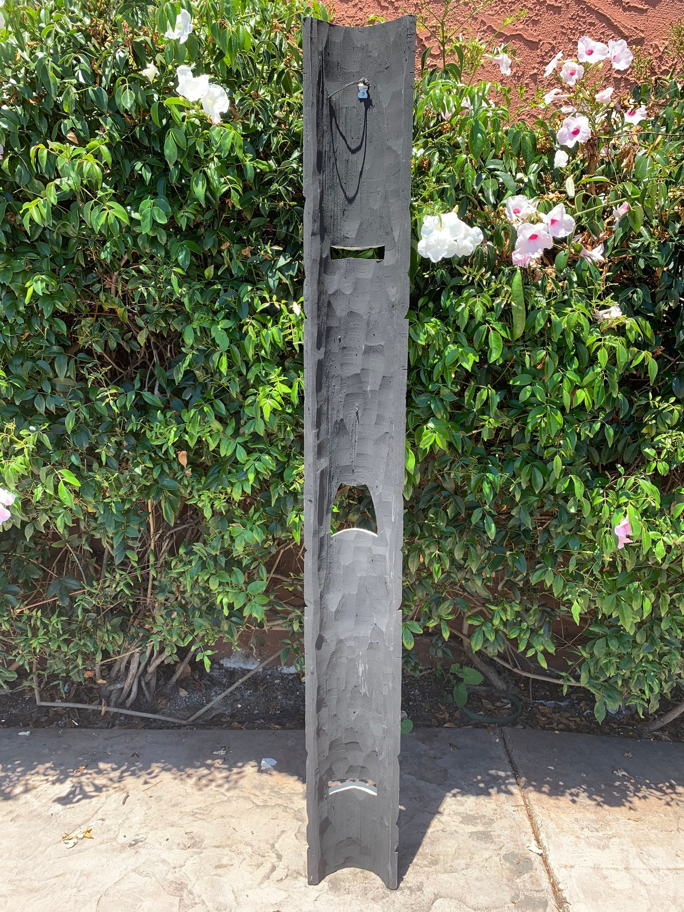 Tall gray wooden sculpture with cutouts in front-yard beside a green hedge and white-pink flowers.