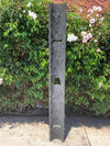 Tall gray wooden sculpture with cutouts in front-yard beside a green hedge and white-pink flowers.