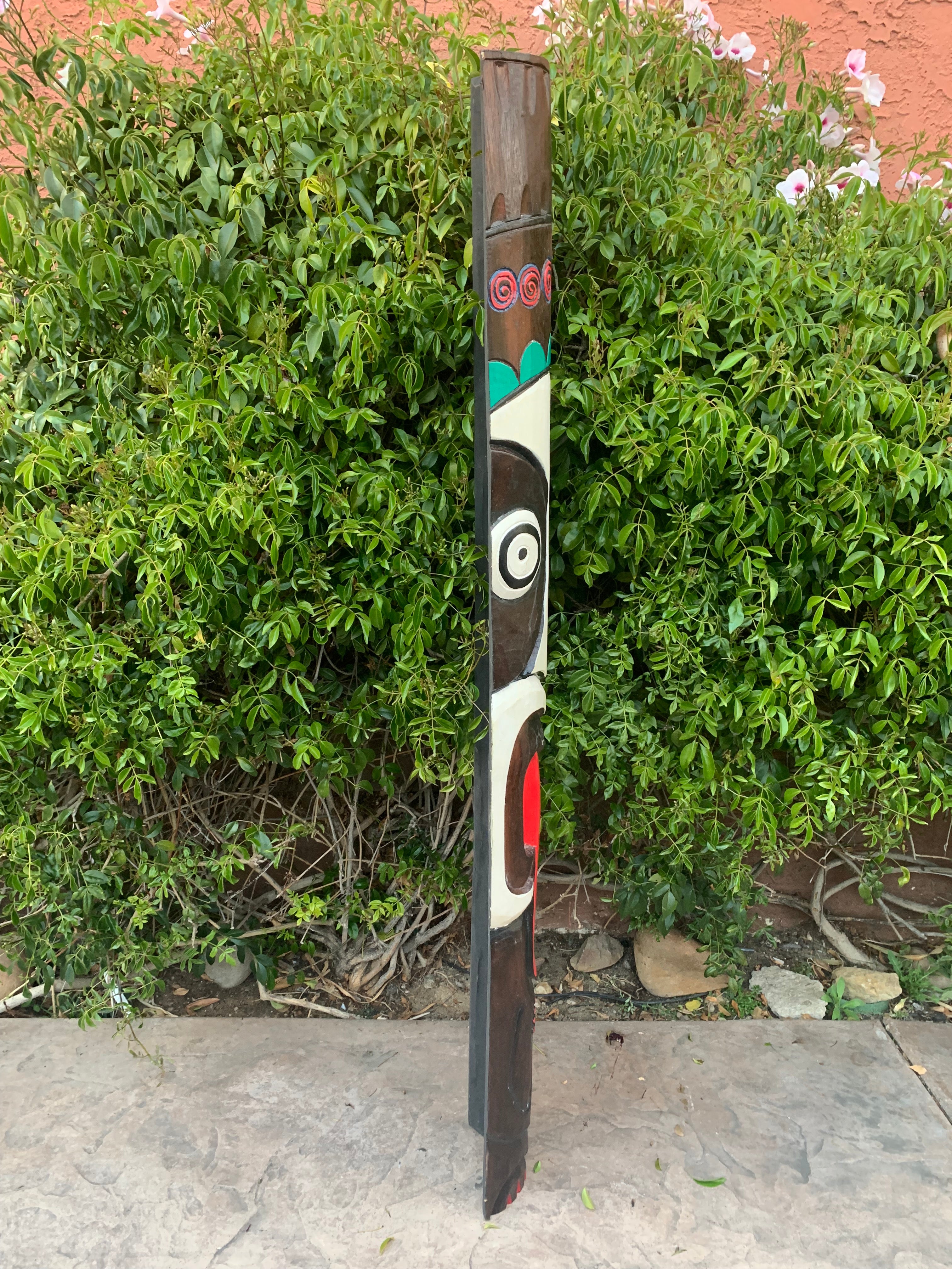 Tall wooden totem pole with a carved face and colorful accents beside green shrubbery