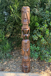 Tall wooden tiki totem pole with carved faces in a garden setting