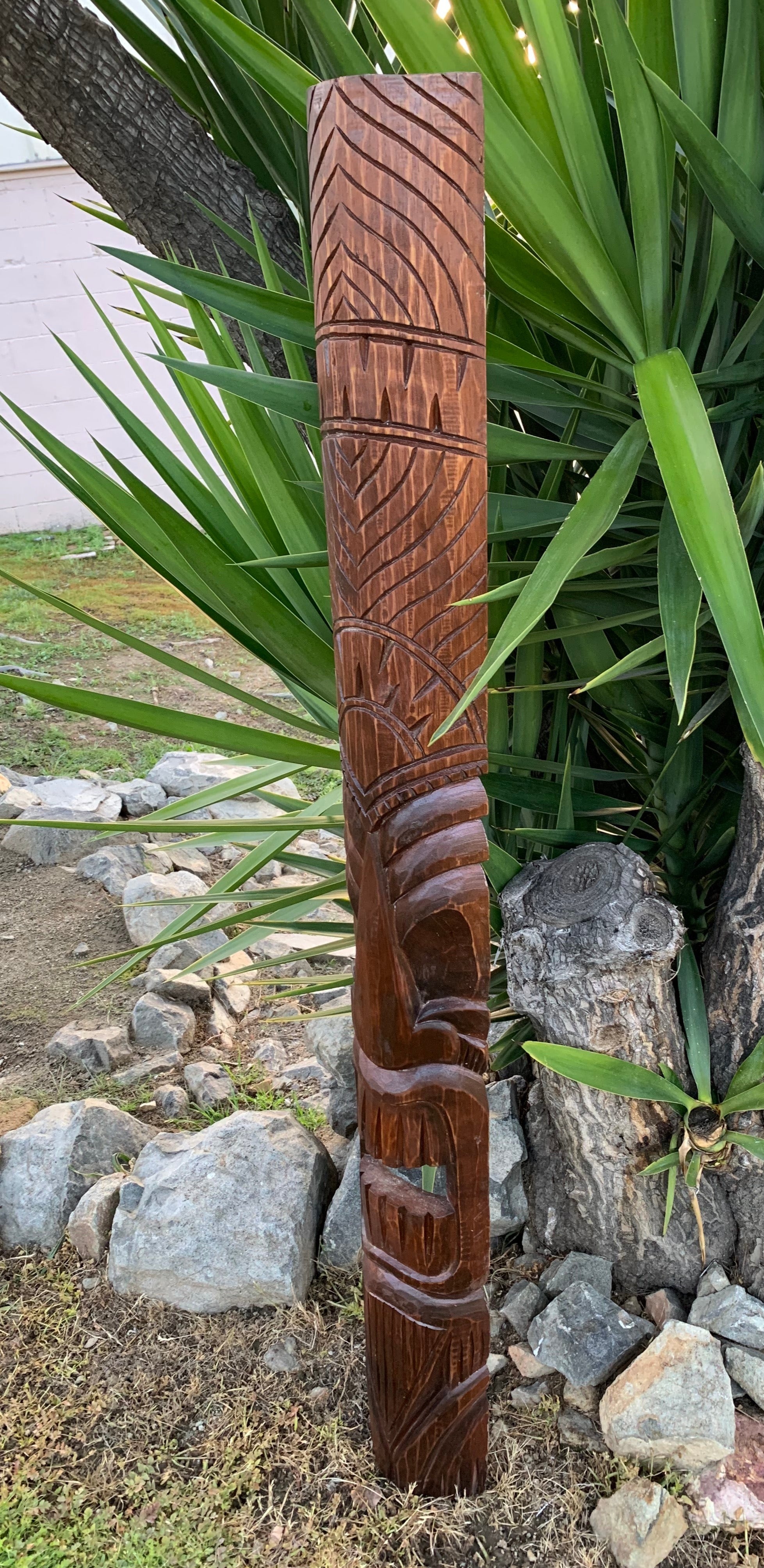 Tall wooden tiki totem carved with geometric patterns outdoors among green plants