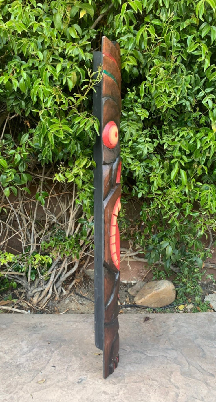 Tall wooden totem sculpture with red circular and curved carved accents outdoors beside green shrubs