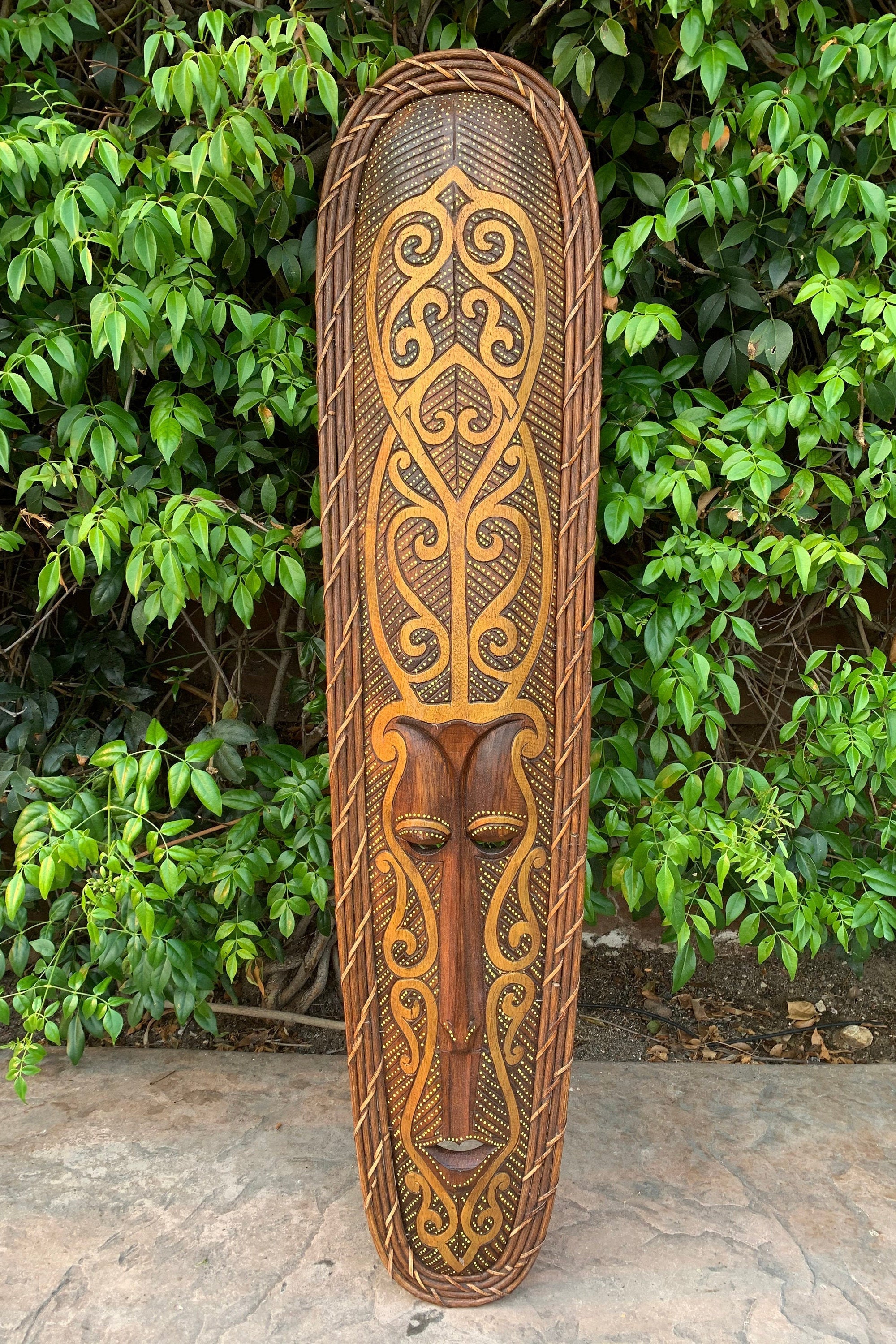 Tall wooden tribal mask with ornate swirling patterns and rope border against green foliage.