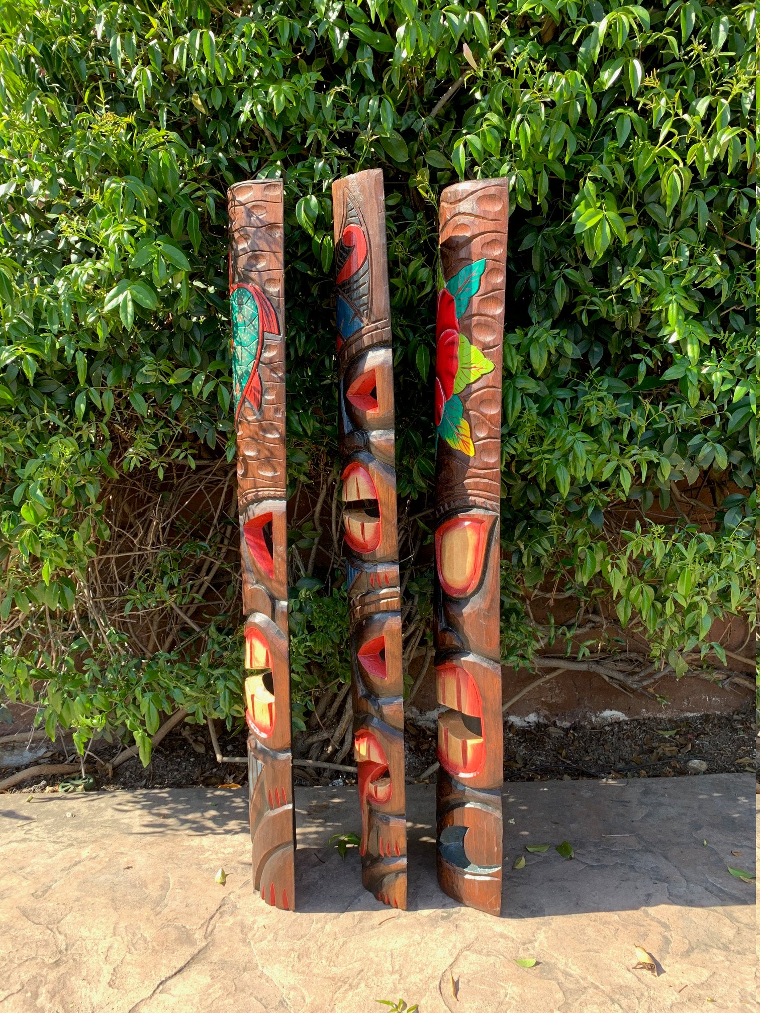 Three tall wooden totem poles with colorful carvings in front of a green leafy hedge.