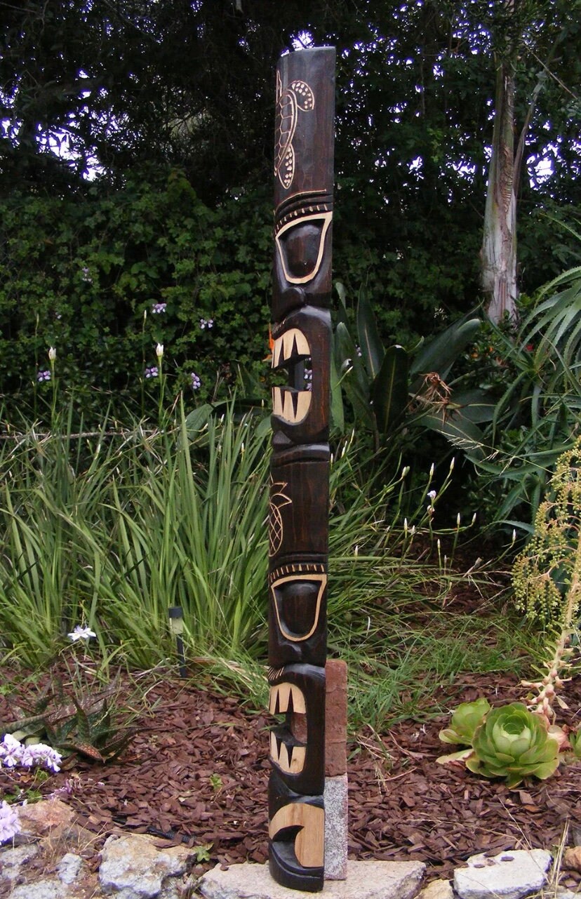 Tall carved wooden totem pole standing in a garden with leafy plants.