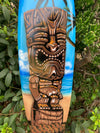 Tiki mask artwork on a blue surfboard leaning against green bushes