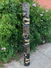 Carved wooden tiki totem pole with multiple faces in a garden of green shrubs and pink hibiscus flowers.