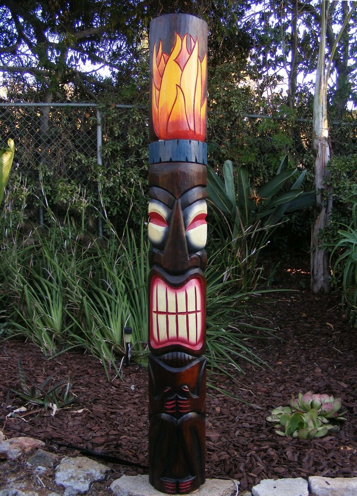 Carved wooden tiki totem pole with a flame-painted top in a garden setting.