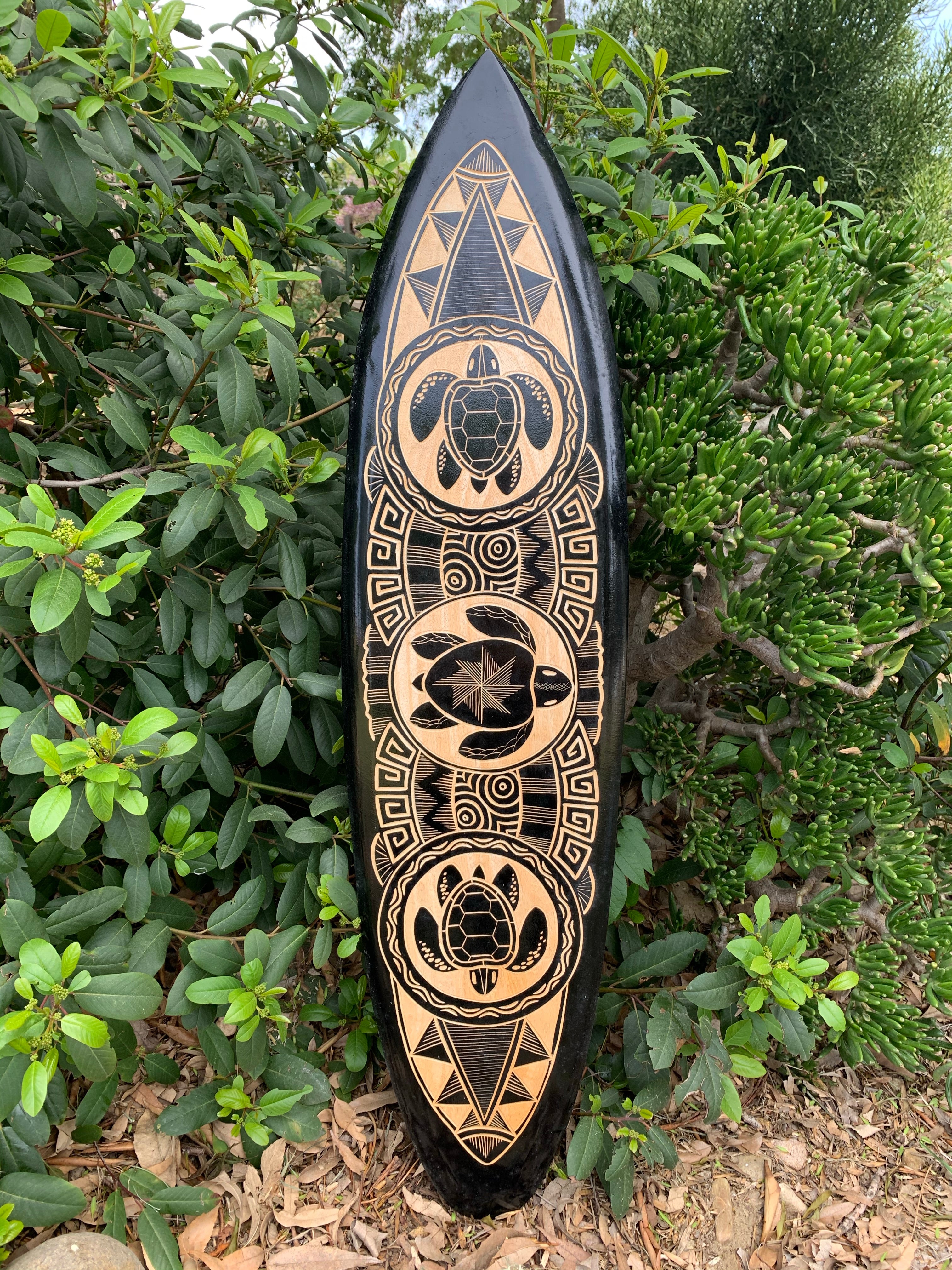 Black surfboard with tan tribal carvings featuring turtles and geometric patterns, leaning against green shrubs.