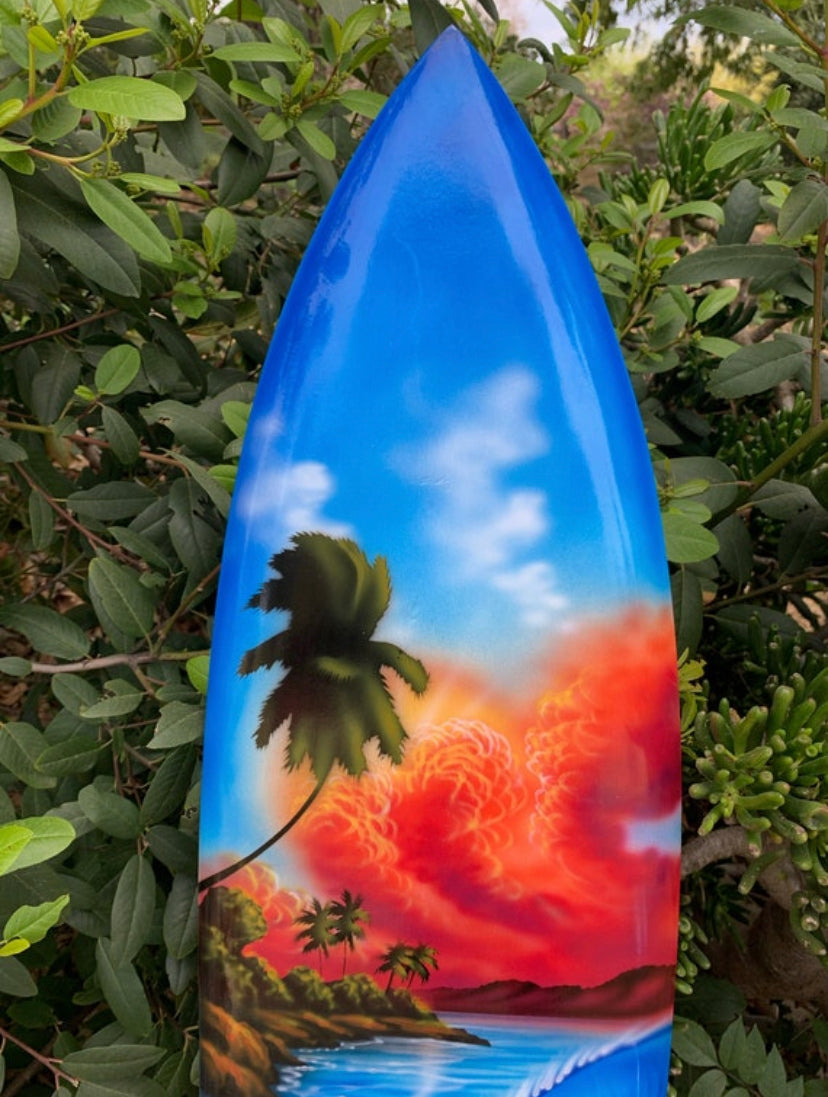 Surfboard with tropical sunset scene, palm trees, blue sky, and orange clouds.