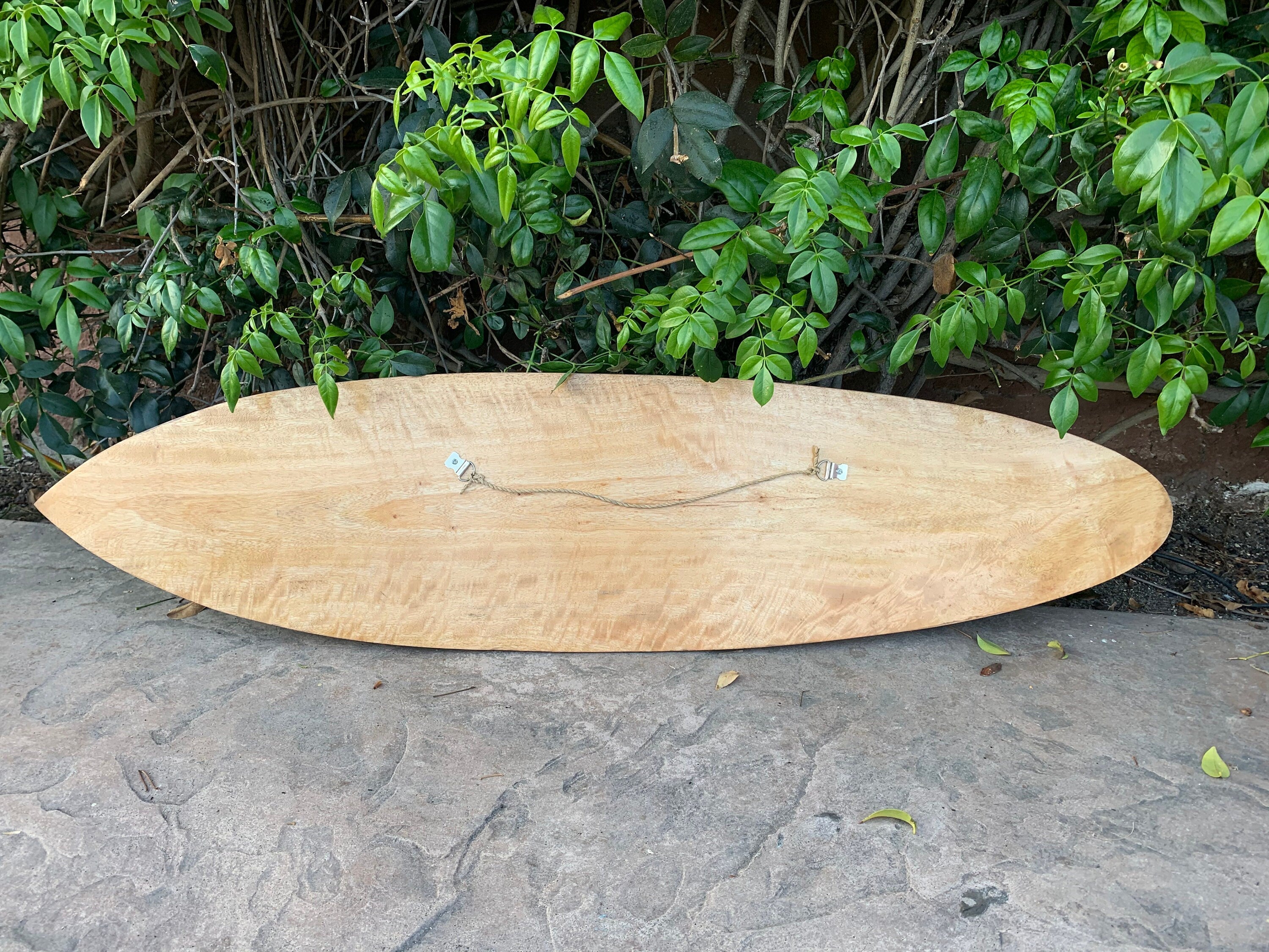 Unfinished wooden surfboard blank on ground with green foliage in the background