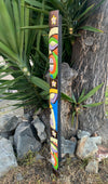 Vertical wooden totem pole with colorful geometric shapes and flower motifs beside a tree