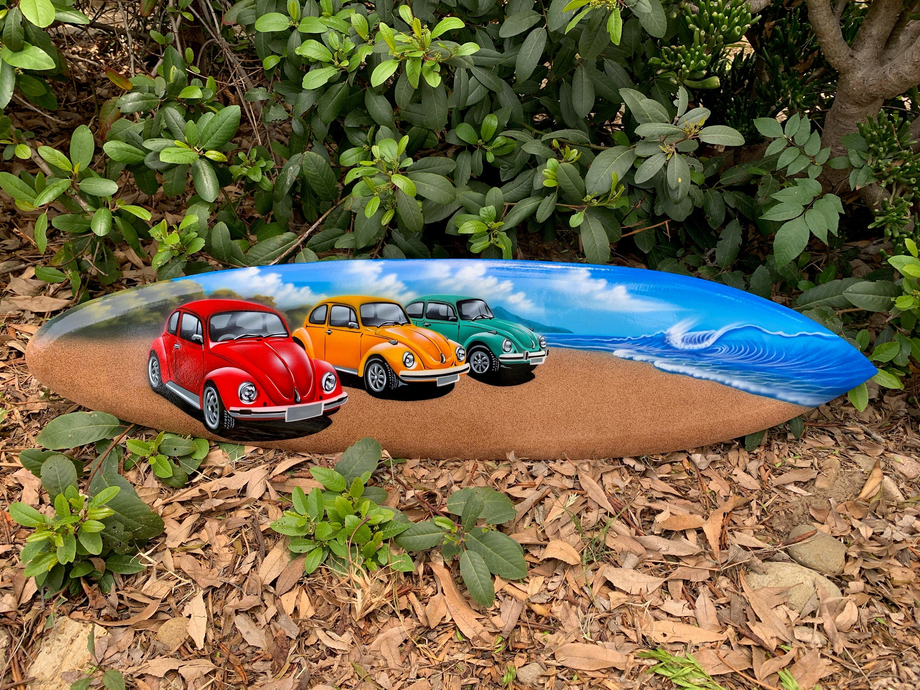 painted-surfboard with red, orange, and green vintage cars on a sandy beach with blue ocean waves
