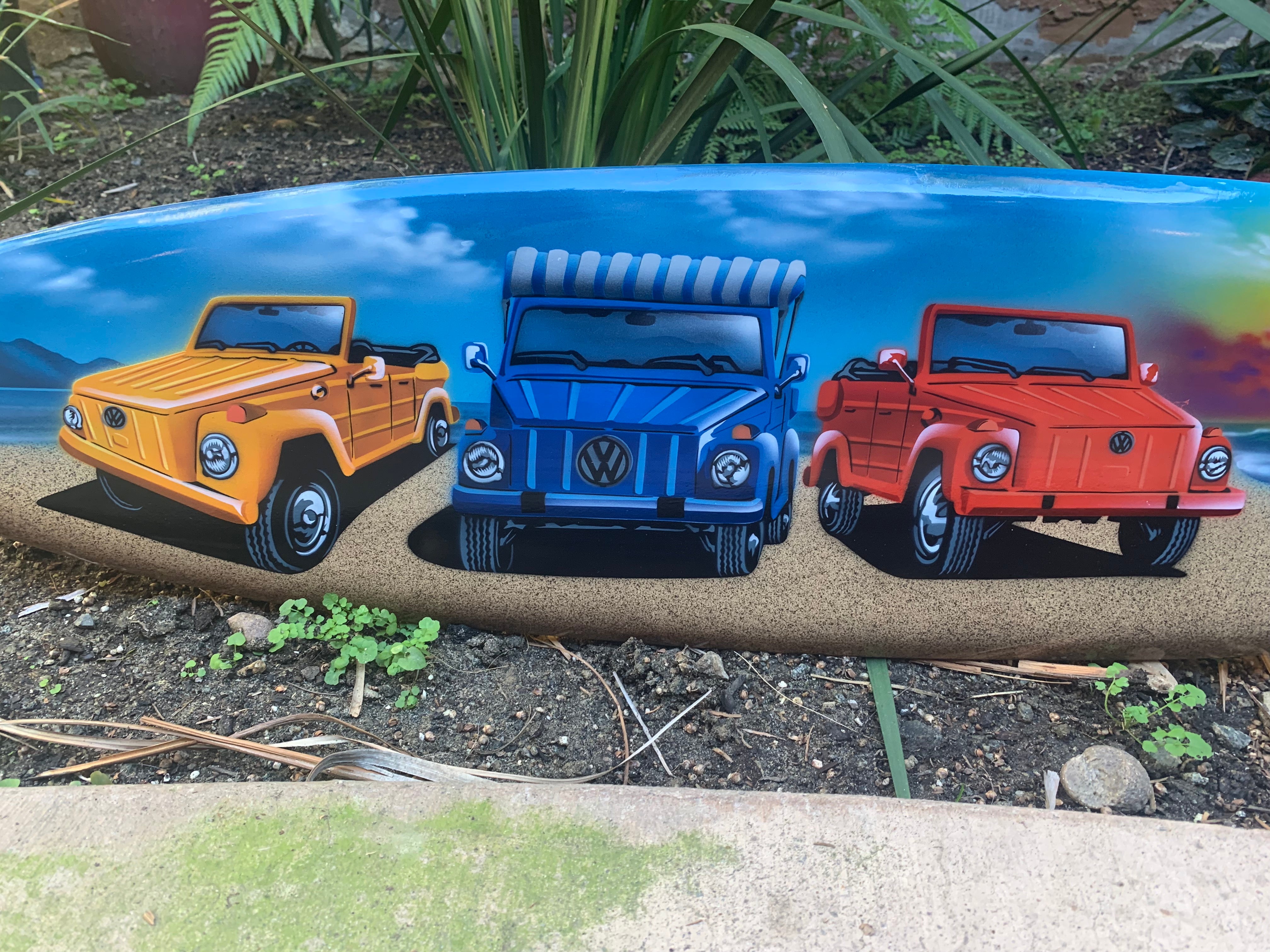 Three colorful VW trucks on a surfboard artwork: yellow, blue, and red.