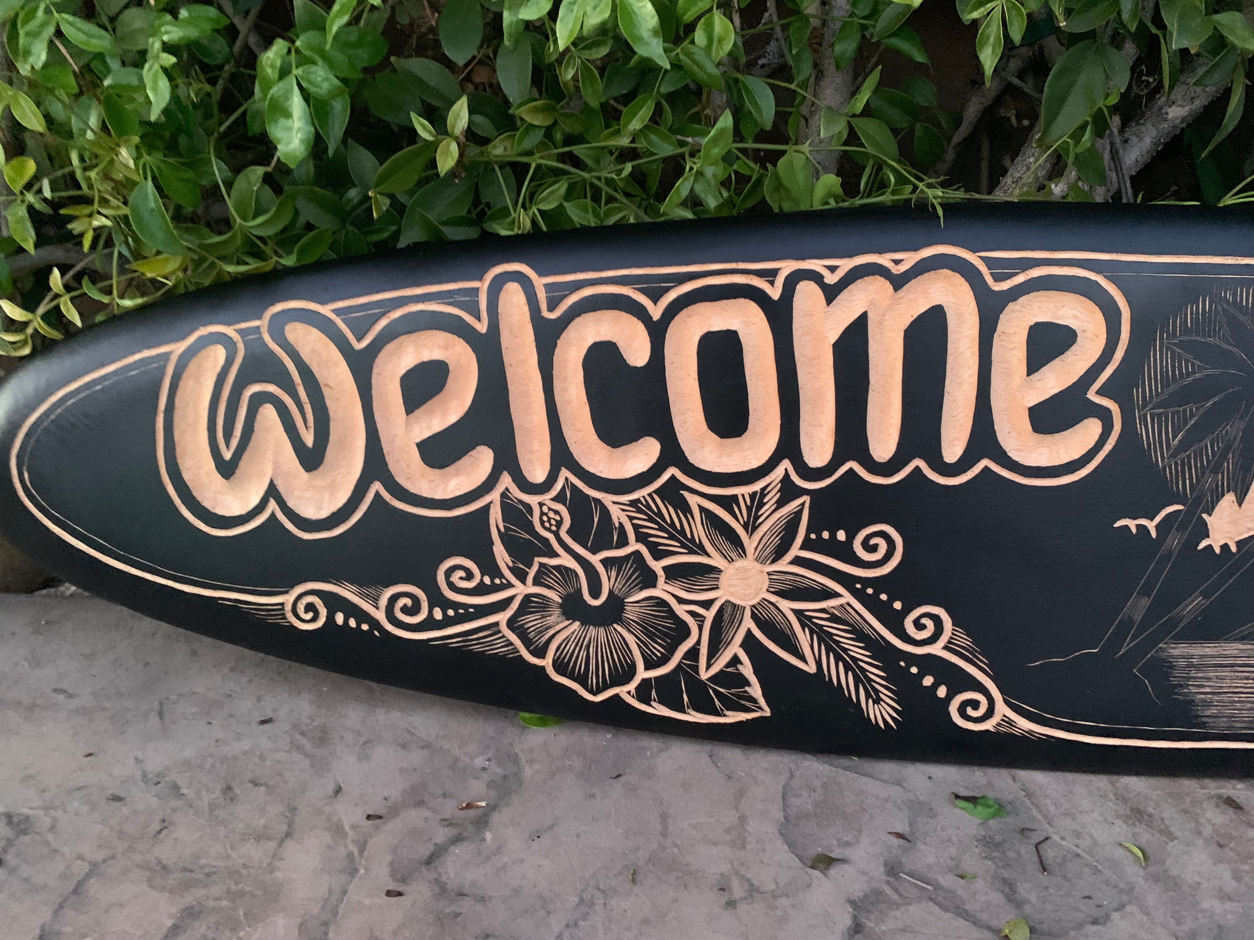 Black surfboard with peach-colored Welcome lettering and decorative floral illustration