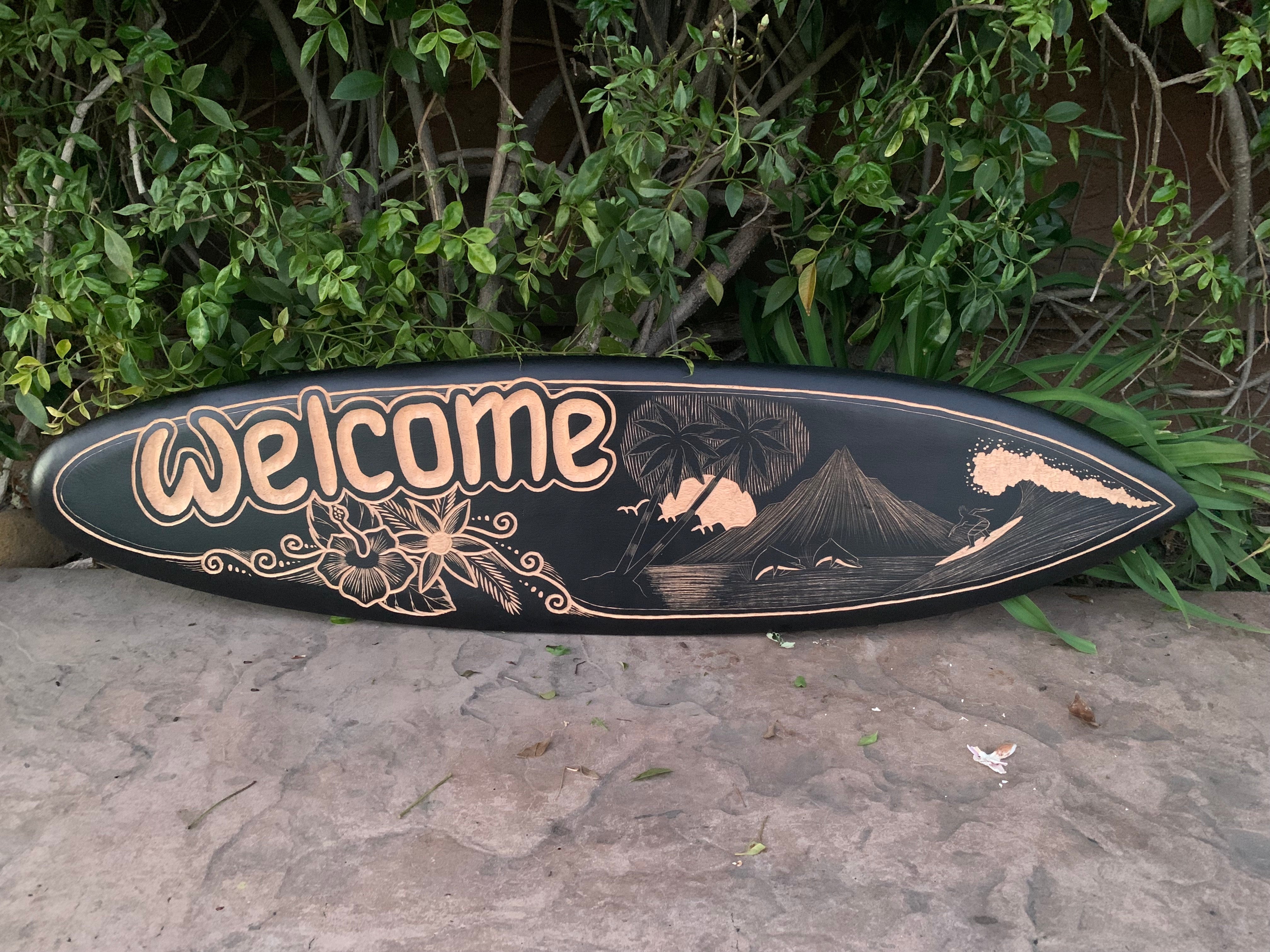 Black surfboard sign with peach-colored Welcome lettering and tropical island scene with palm trees and a volcano