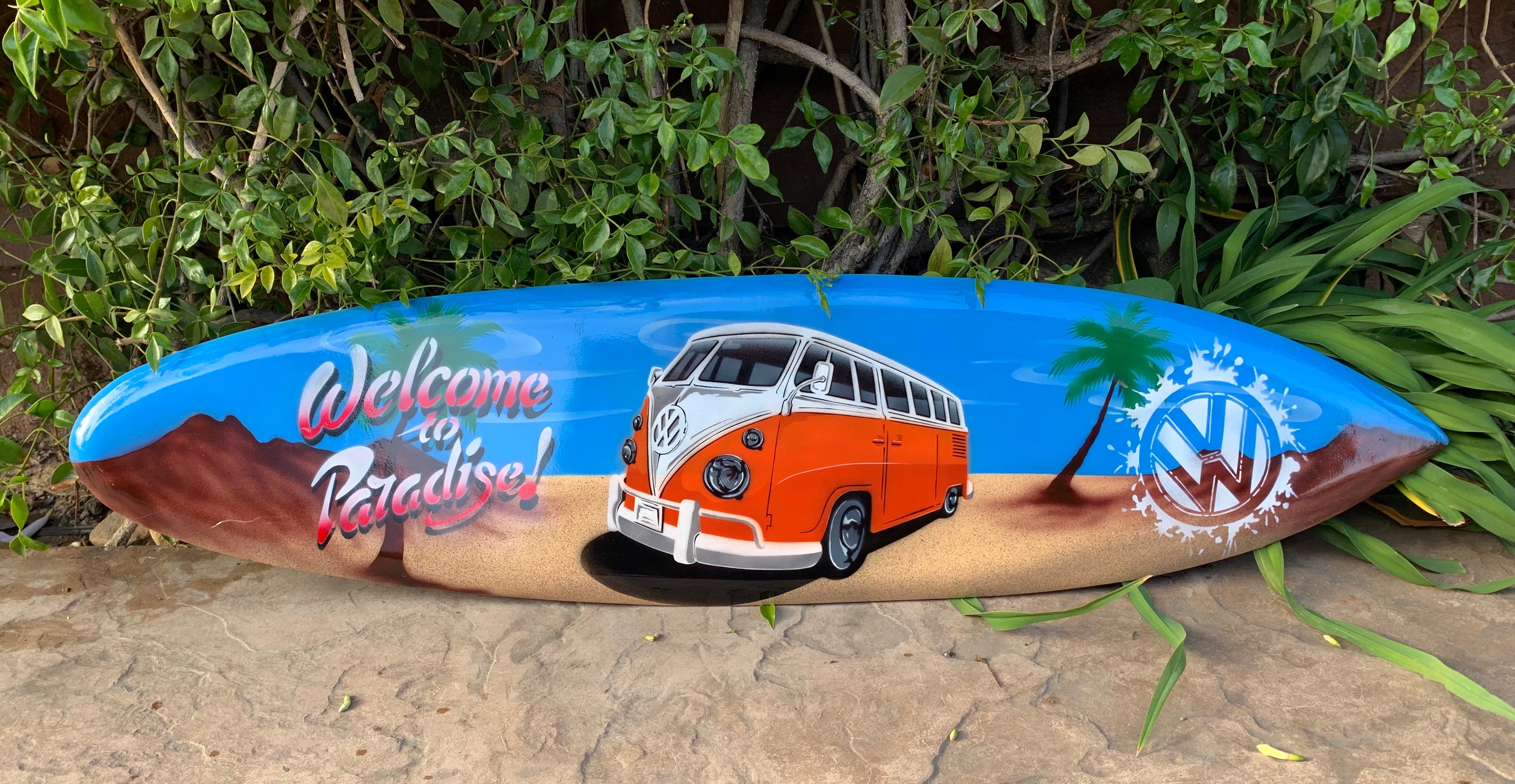 Colorful painted surfboard with orange VW bus, palm trees and 'Welcome to Paradise' text on a beach scene