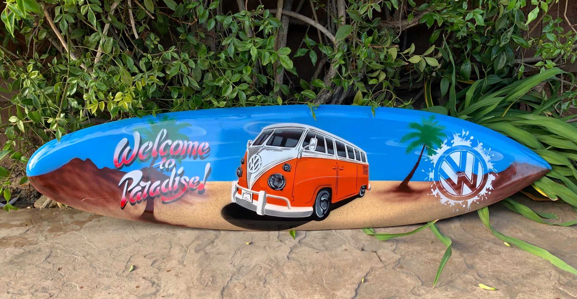 Colorful painted surfboard with orange VW bus, palm trees and 'Welcome to Paradise' text on a beach scene