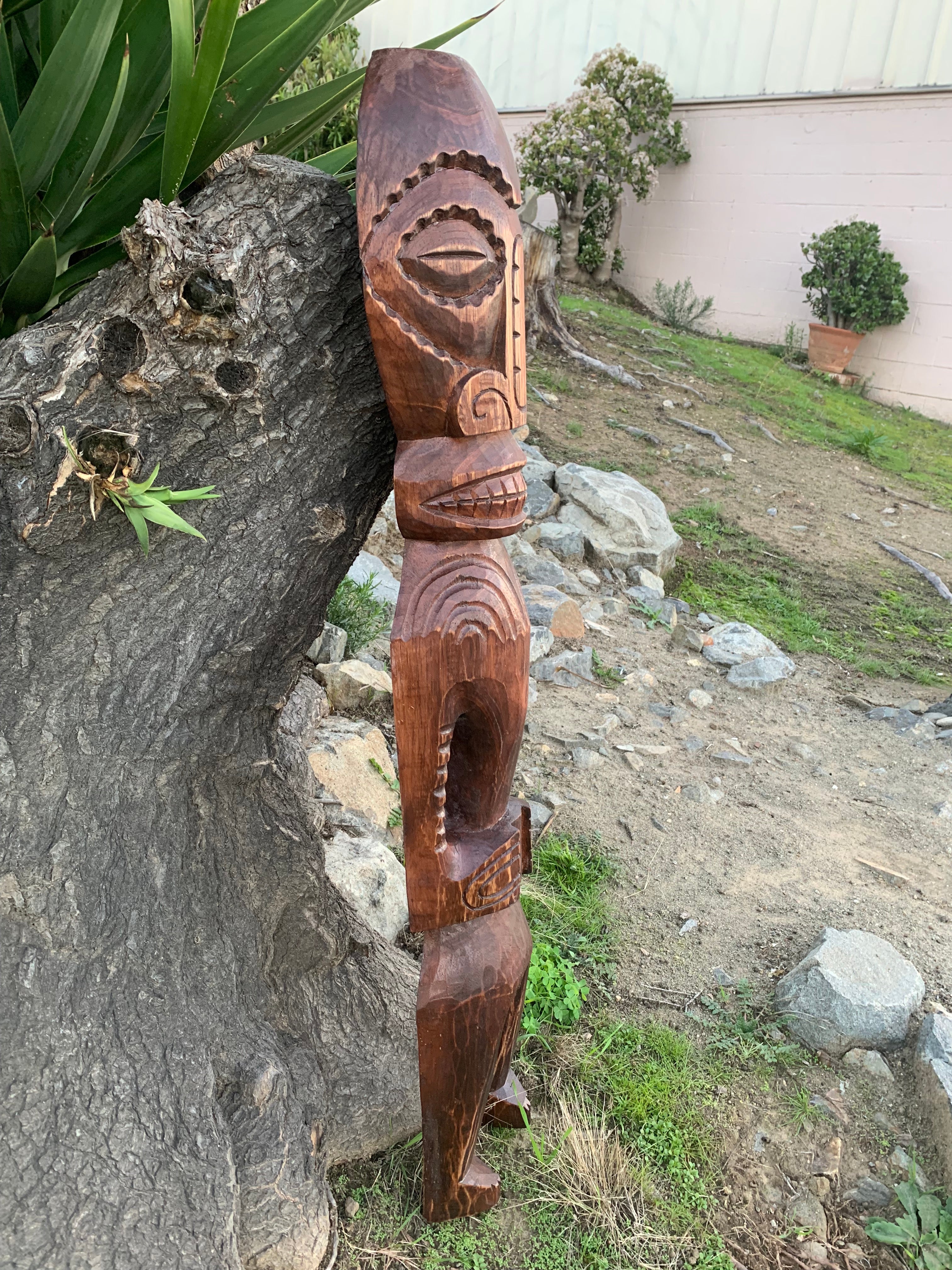 Tall wooden carved totem pole leaning against a tree in a garden with rocks and potted plants.