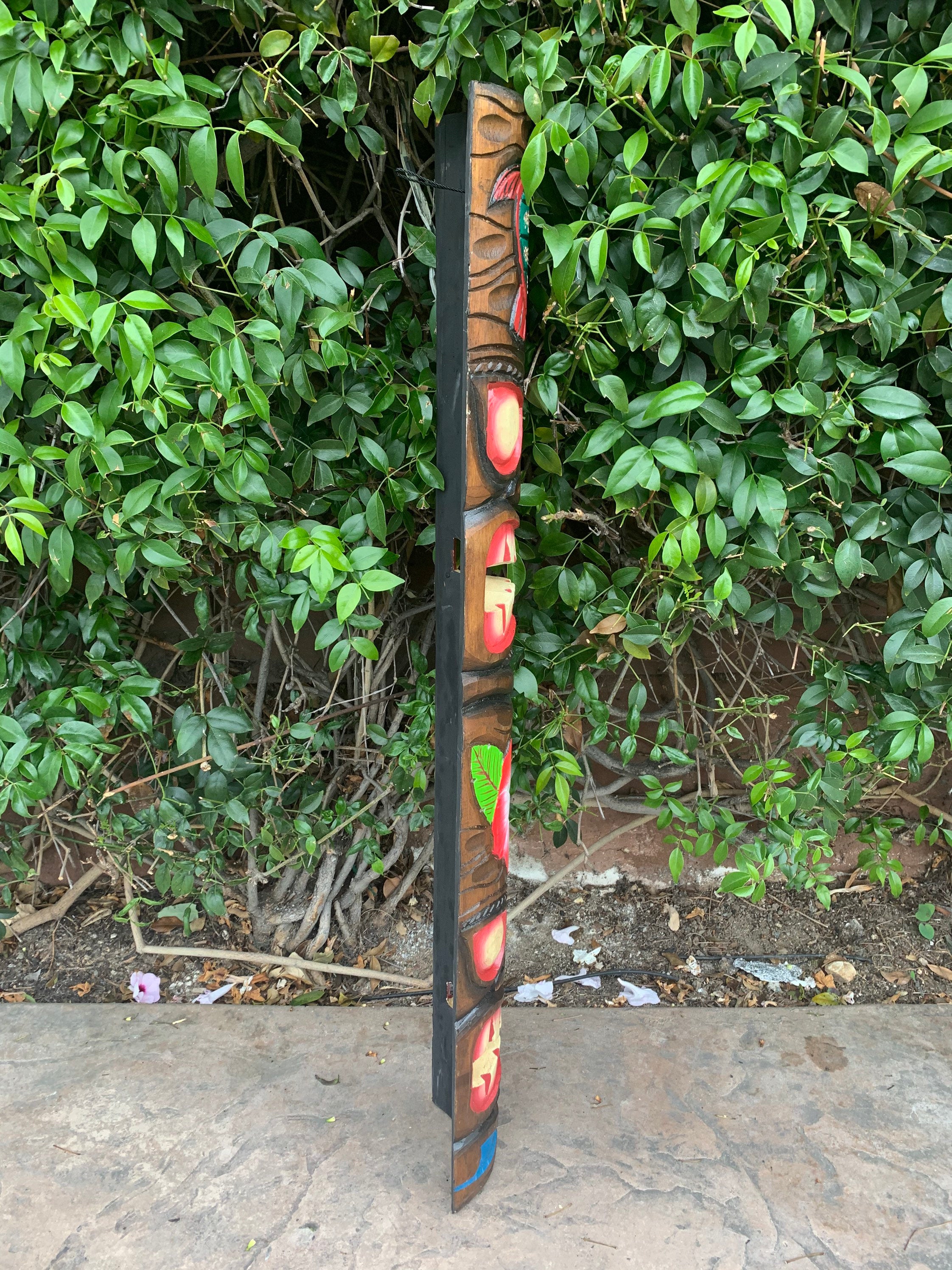 Carved wooden totem pole with red and yellow faces leaning beside a green hedge.