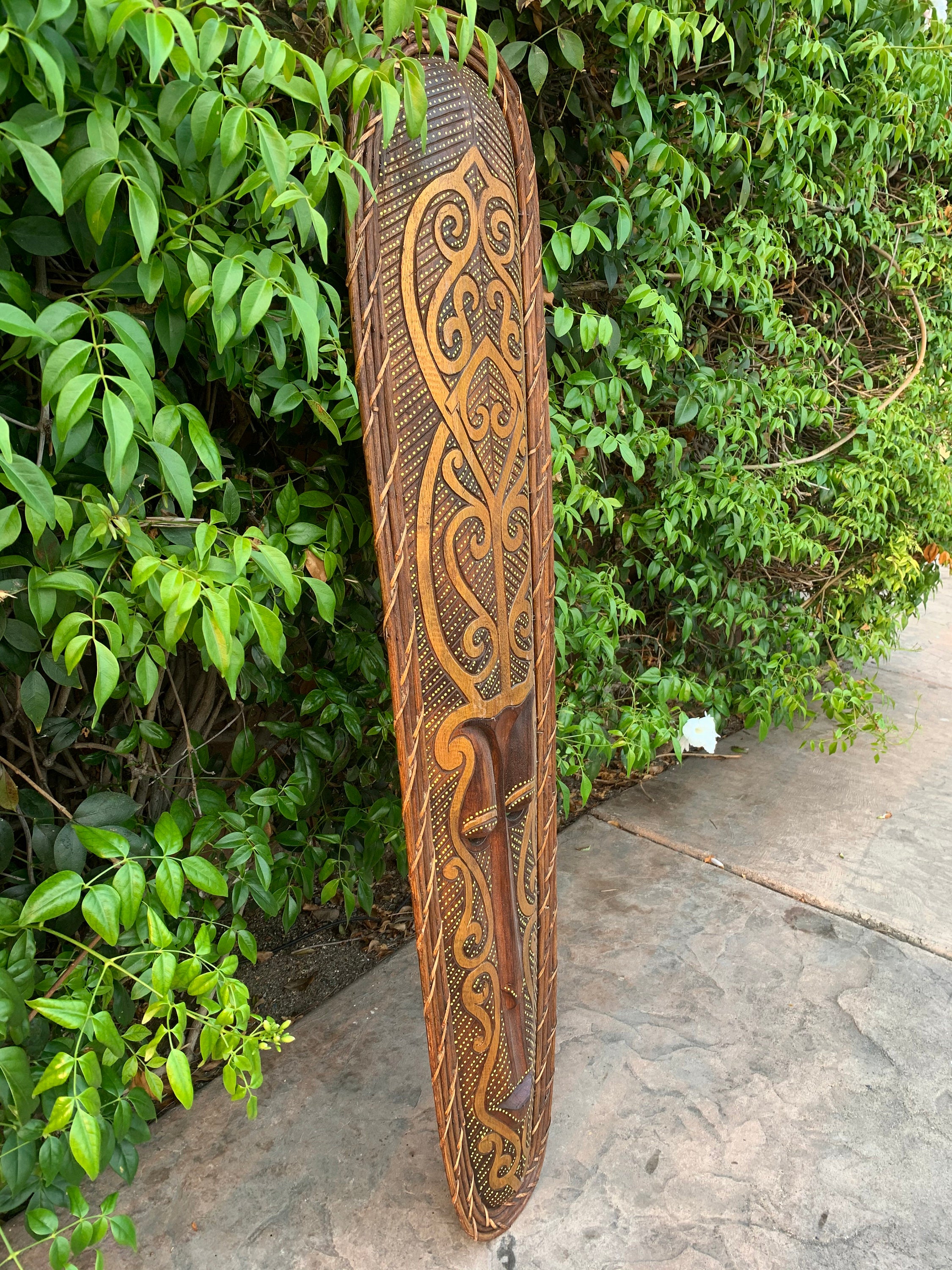 Wooden carved tribal panel with spiral motifs leaning against a green hedge.