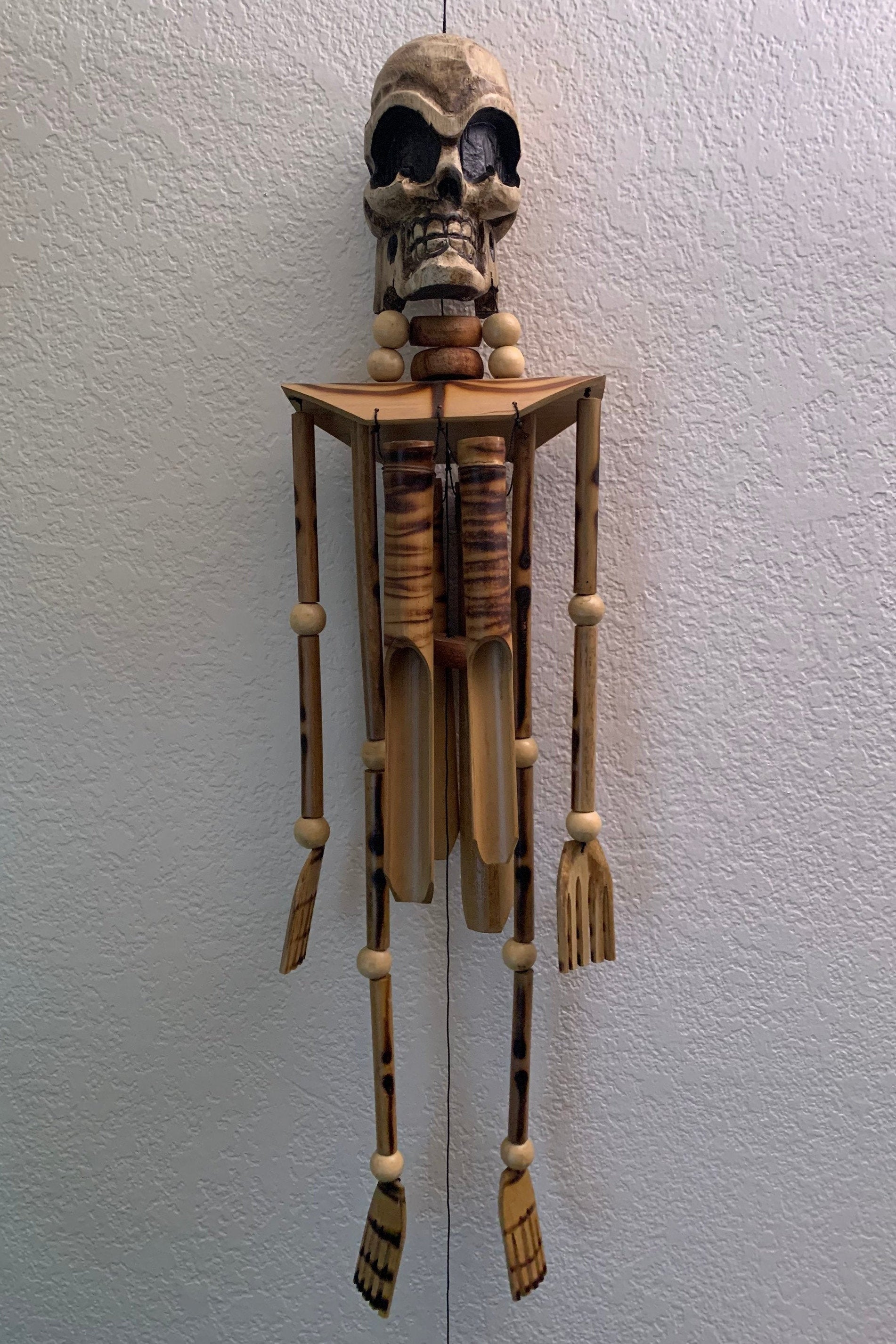 Wooden skeletal marionette with carved skull and bead necklace against a textured wall.