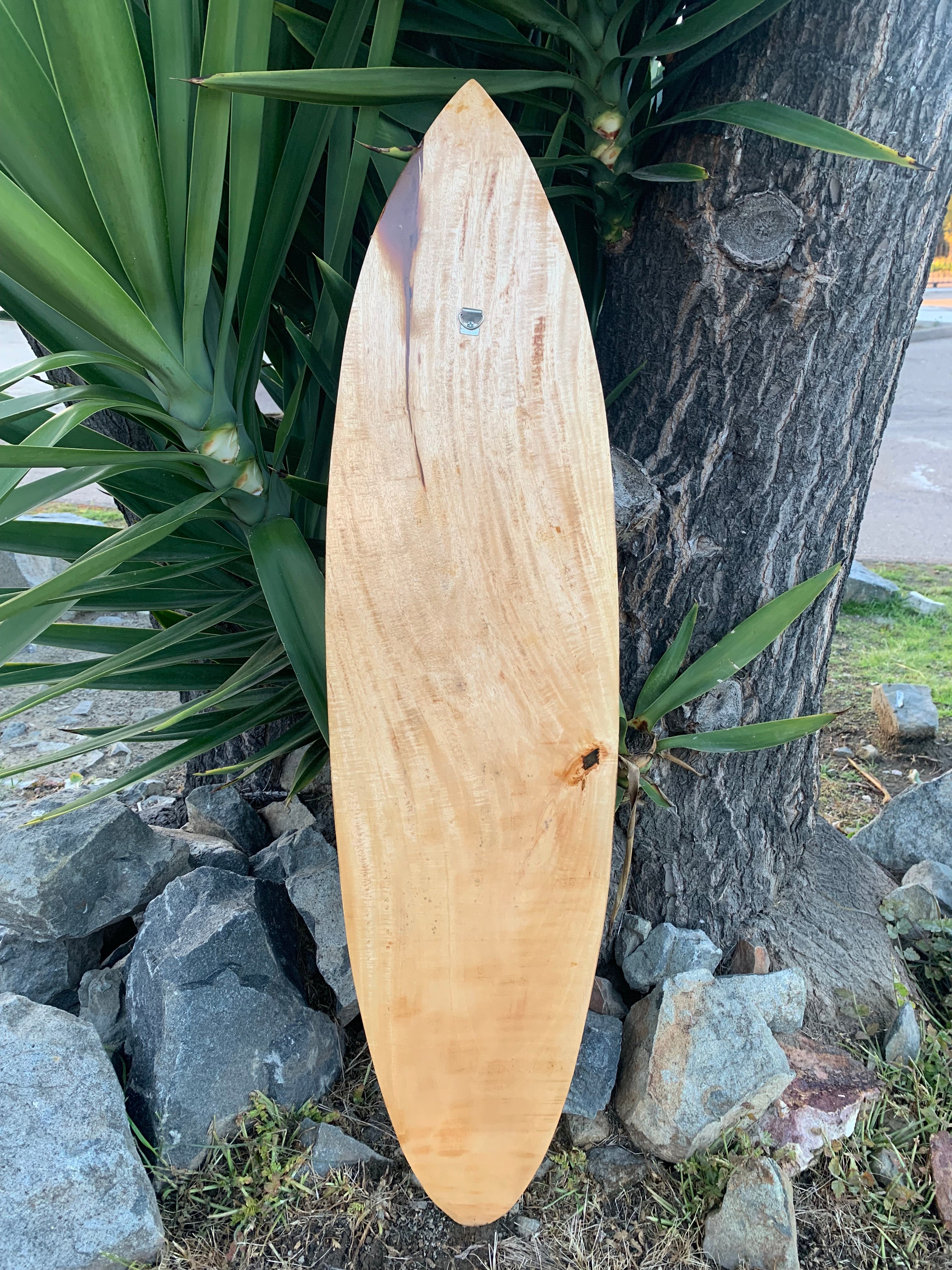 Natural wood surfboard leaning against a tree with greenery and rocks nearby