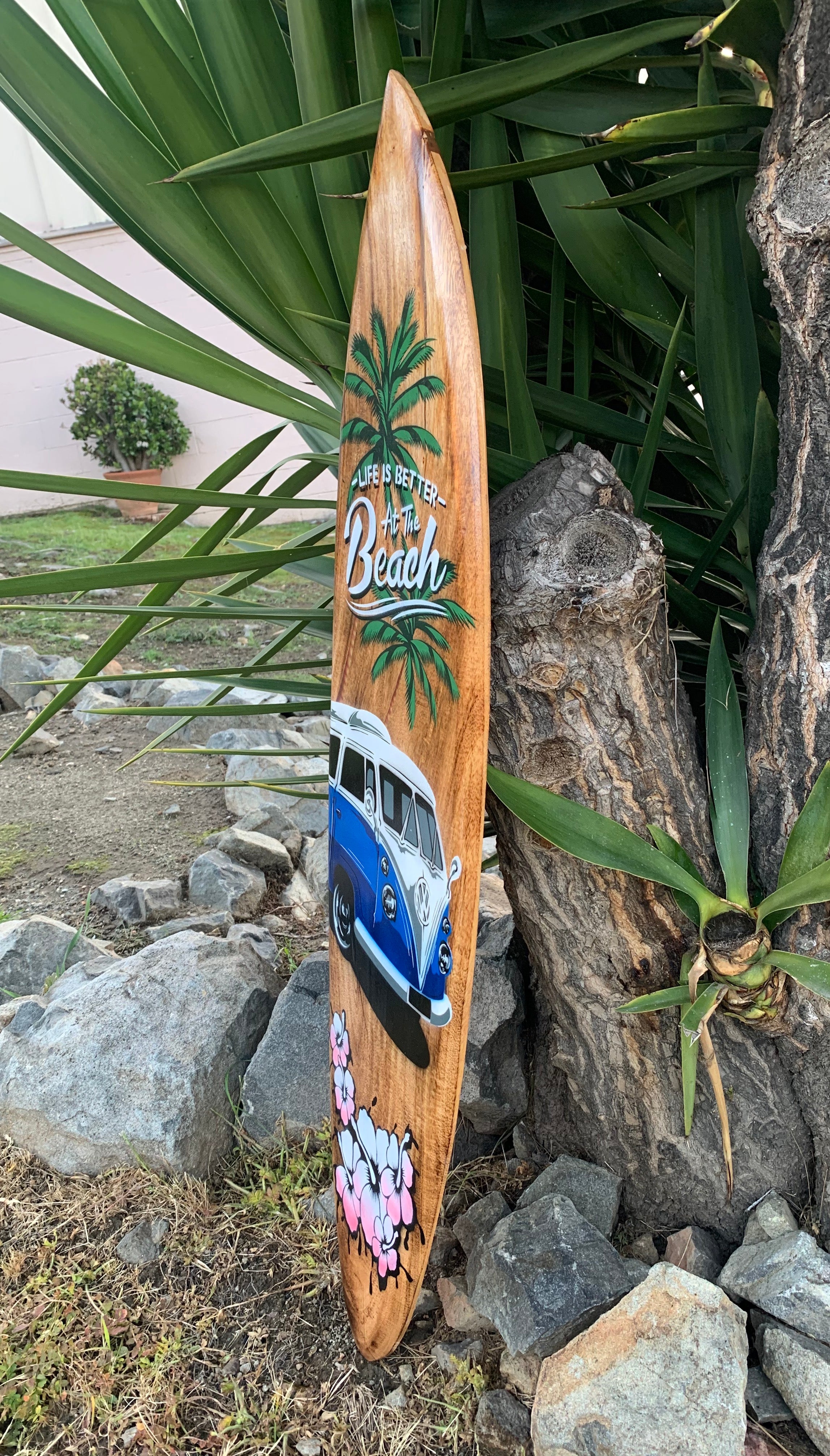 Wooden surfboard wall art with palm trees, a blue VW bus, and pink hibiscus flowers.