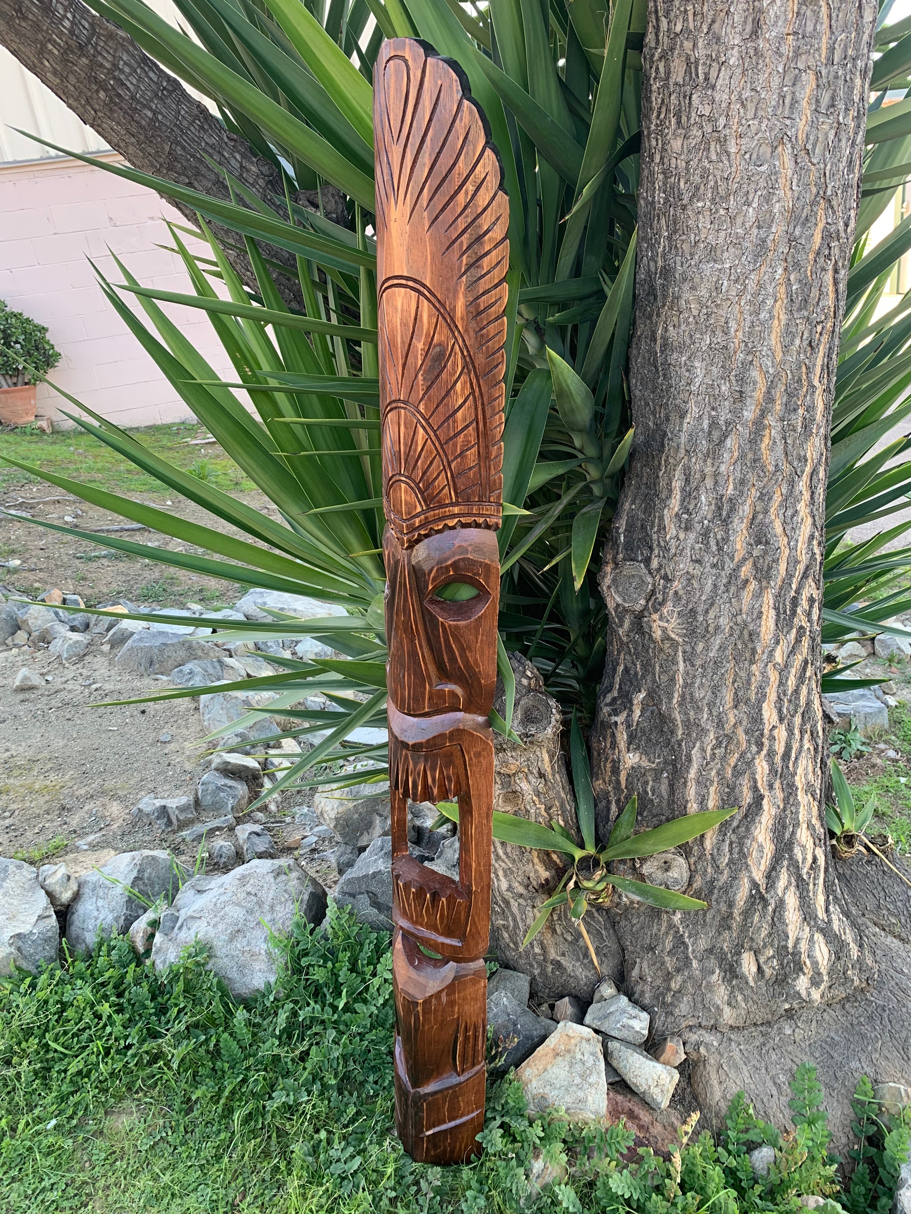 Wooden tiki carving with feathered headdress leaning against a tree in a garden