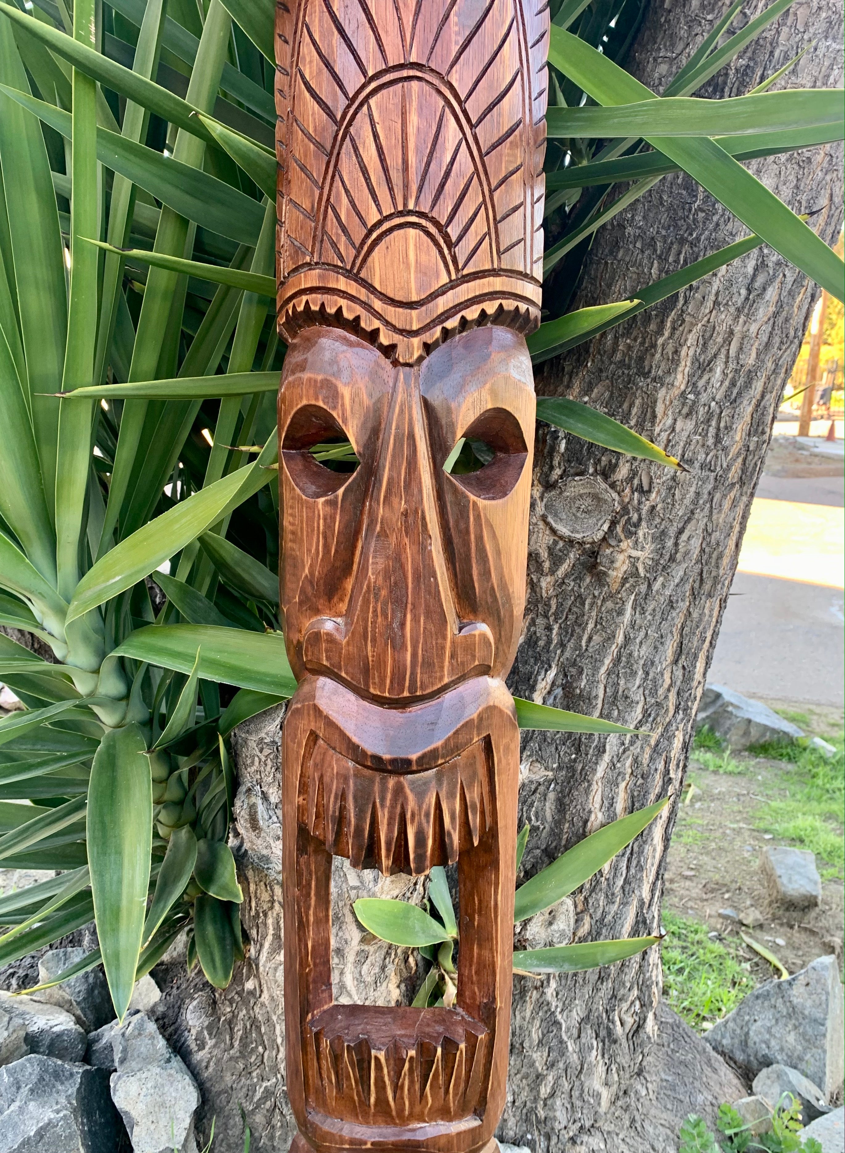Wooden carved tiki mask with sunburst headdress outdoors beside a tree