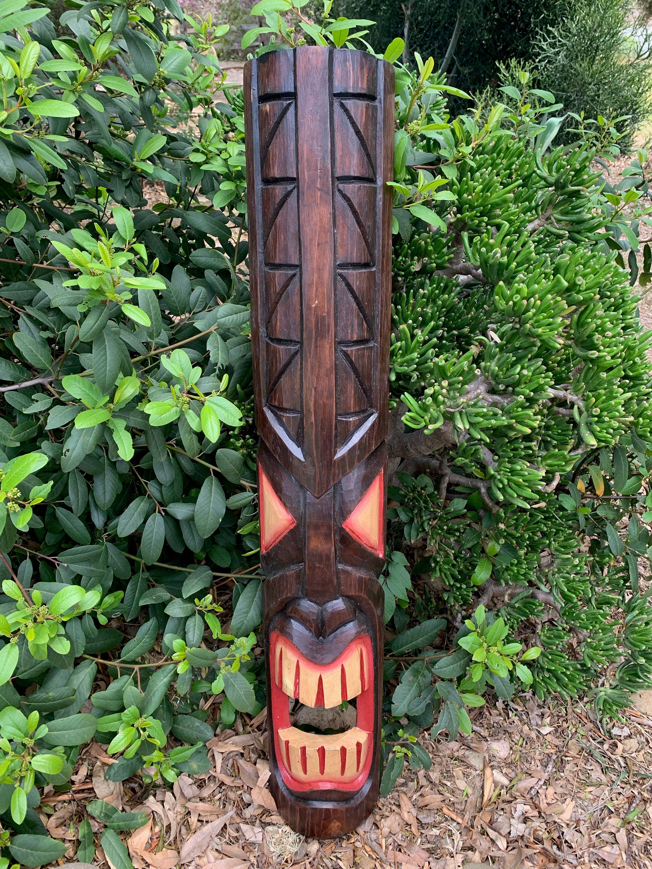 tall carved wooden tiki mask statue outdoors among green shrubs