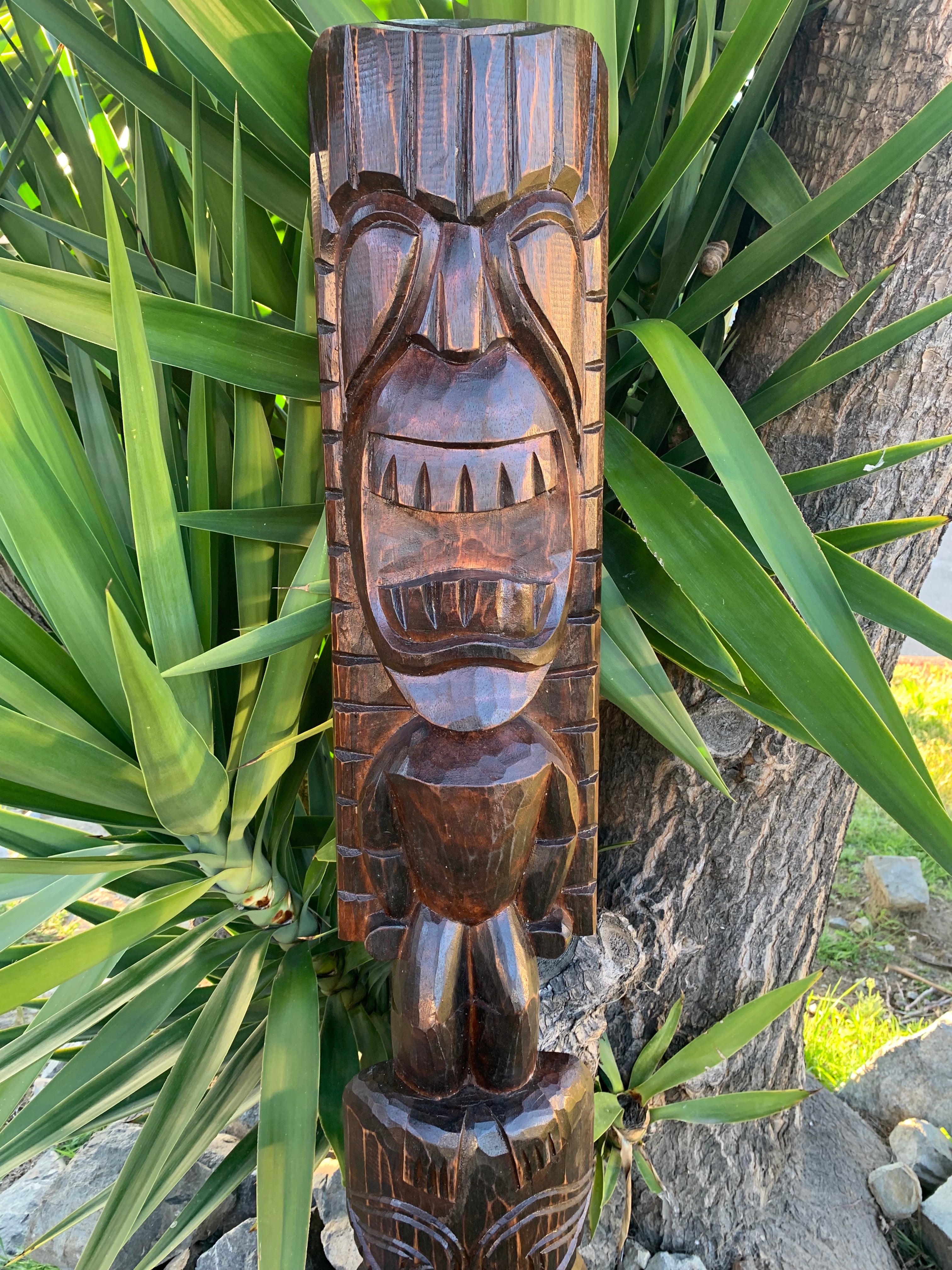 carved wooden tiki statue with a wide toothy grin outdoors among green plants