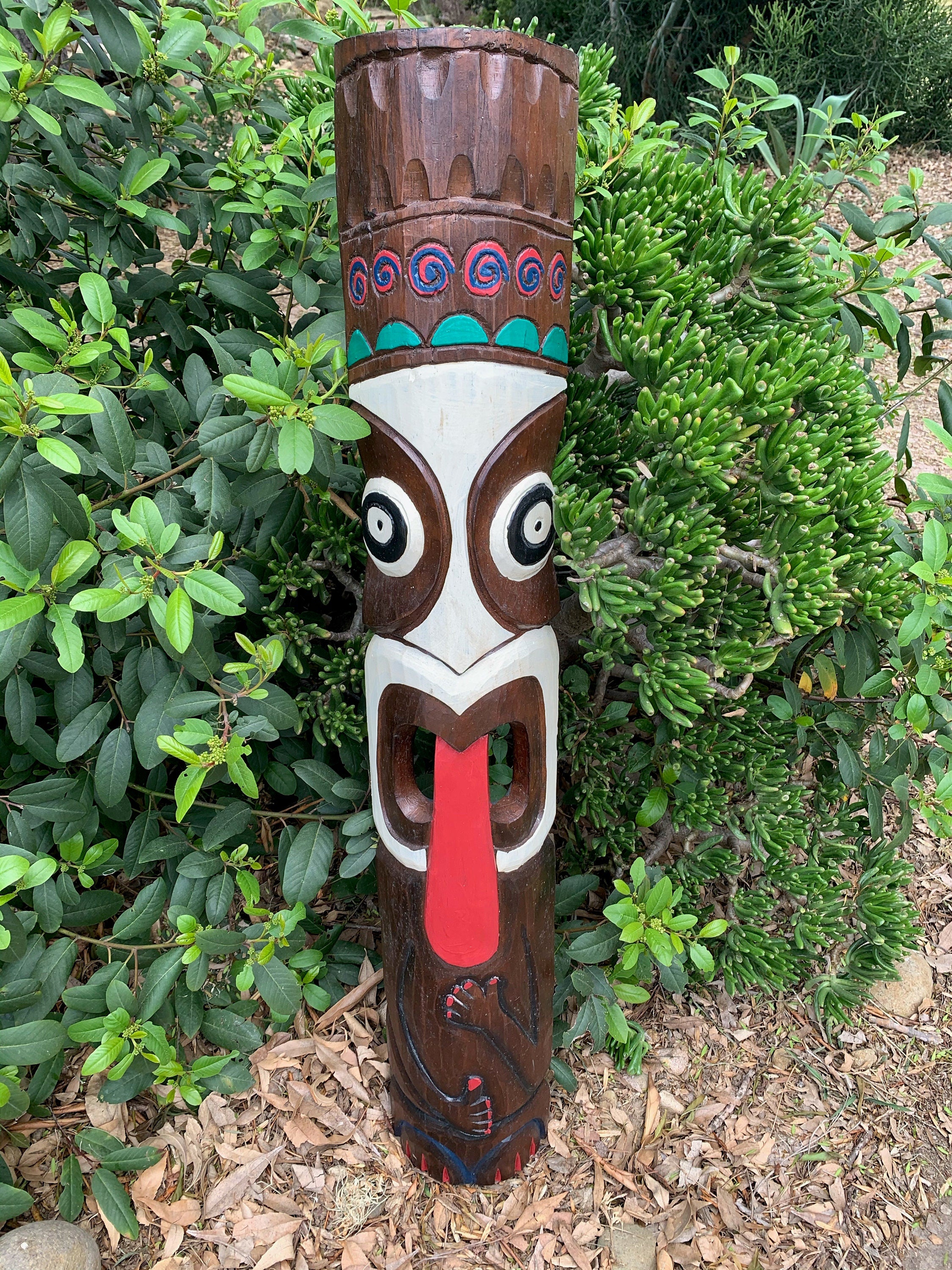 Carved wooden tiki statue with turquoise headband and red tongue in a garden