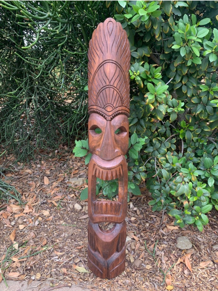 Carved wooden tiki statue with tall headdress and open mouth, set among greenery.