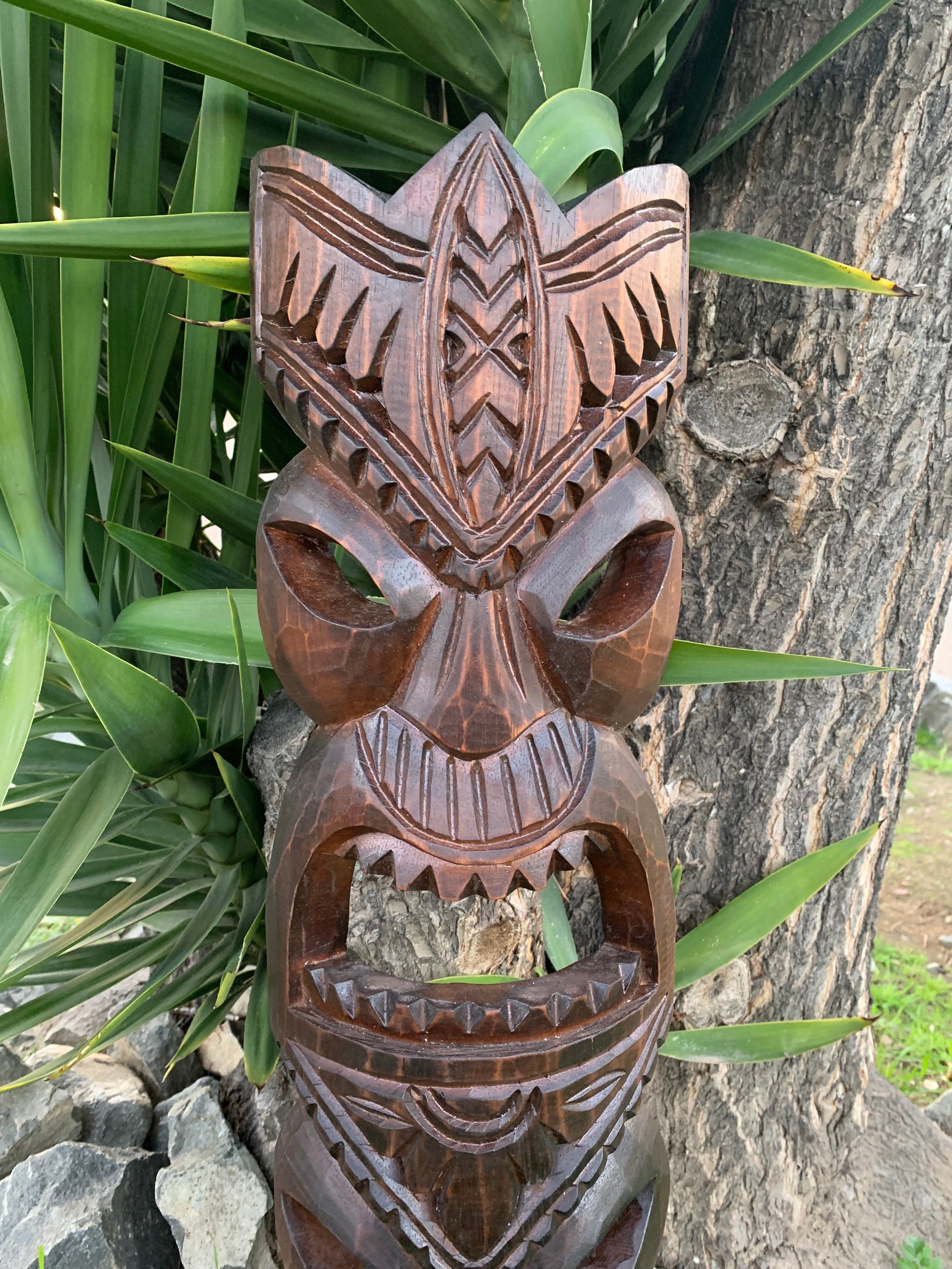 Carved wooden tiki statue with geometric patterns outdoors beside a tree and green leaves.