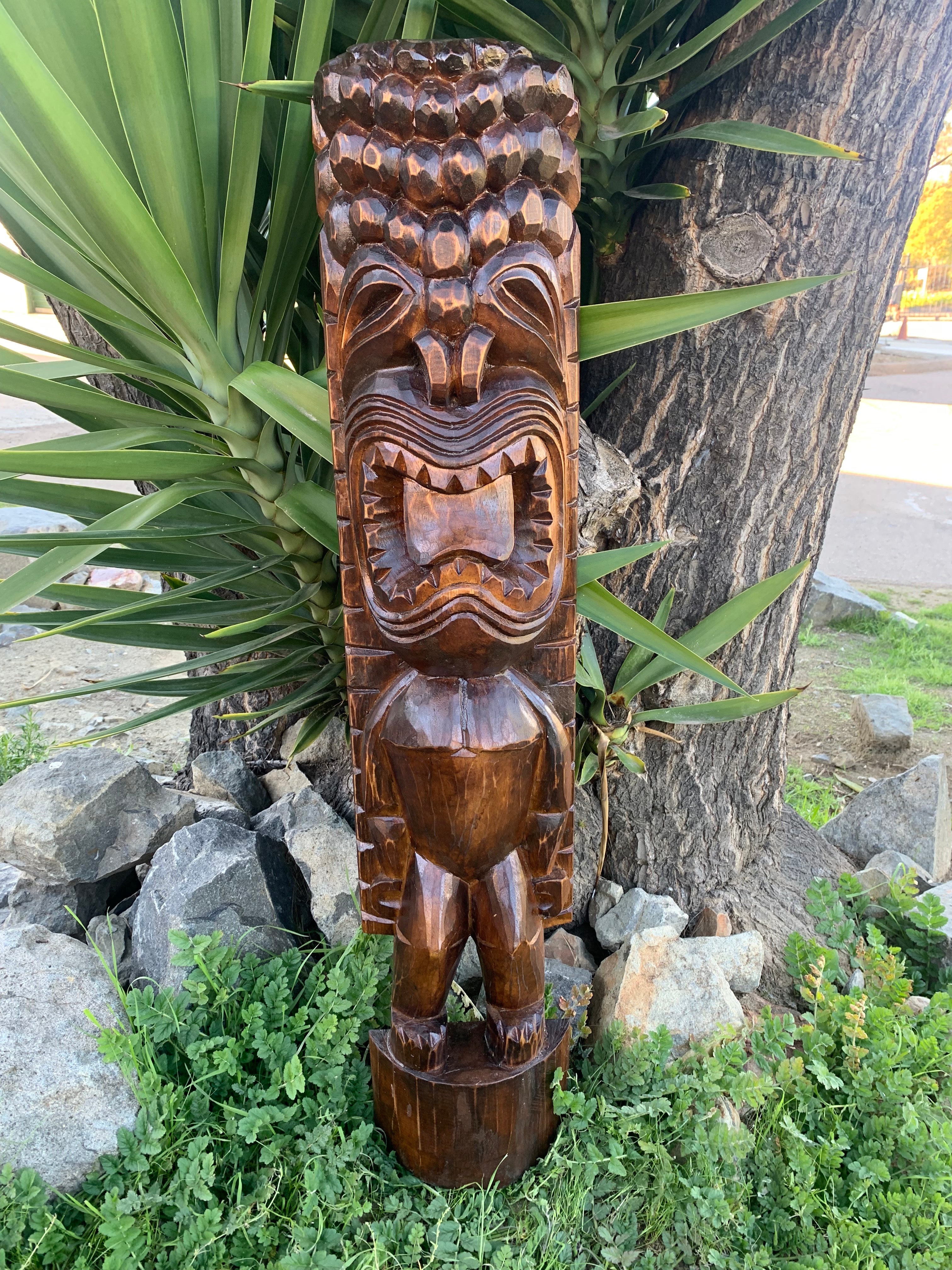 Wooden tiki statue carved with a fierce face, wide-open mouth, and geometric detailing outdoors