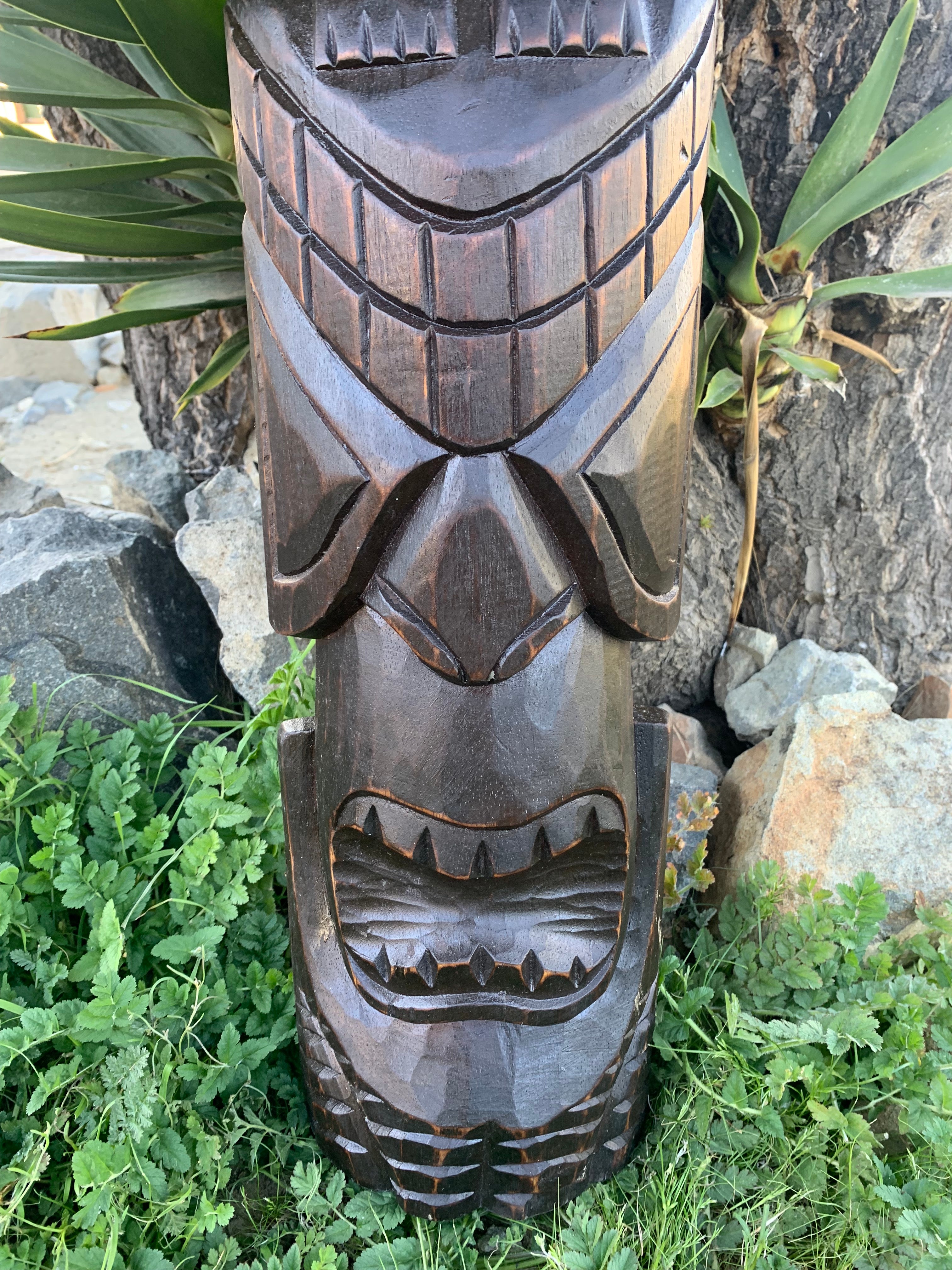 Carved wooden tiki totem pole with grinning faces outdoors among rocks and greenery