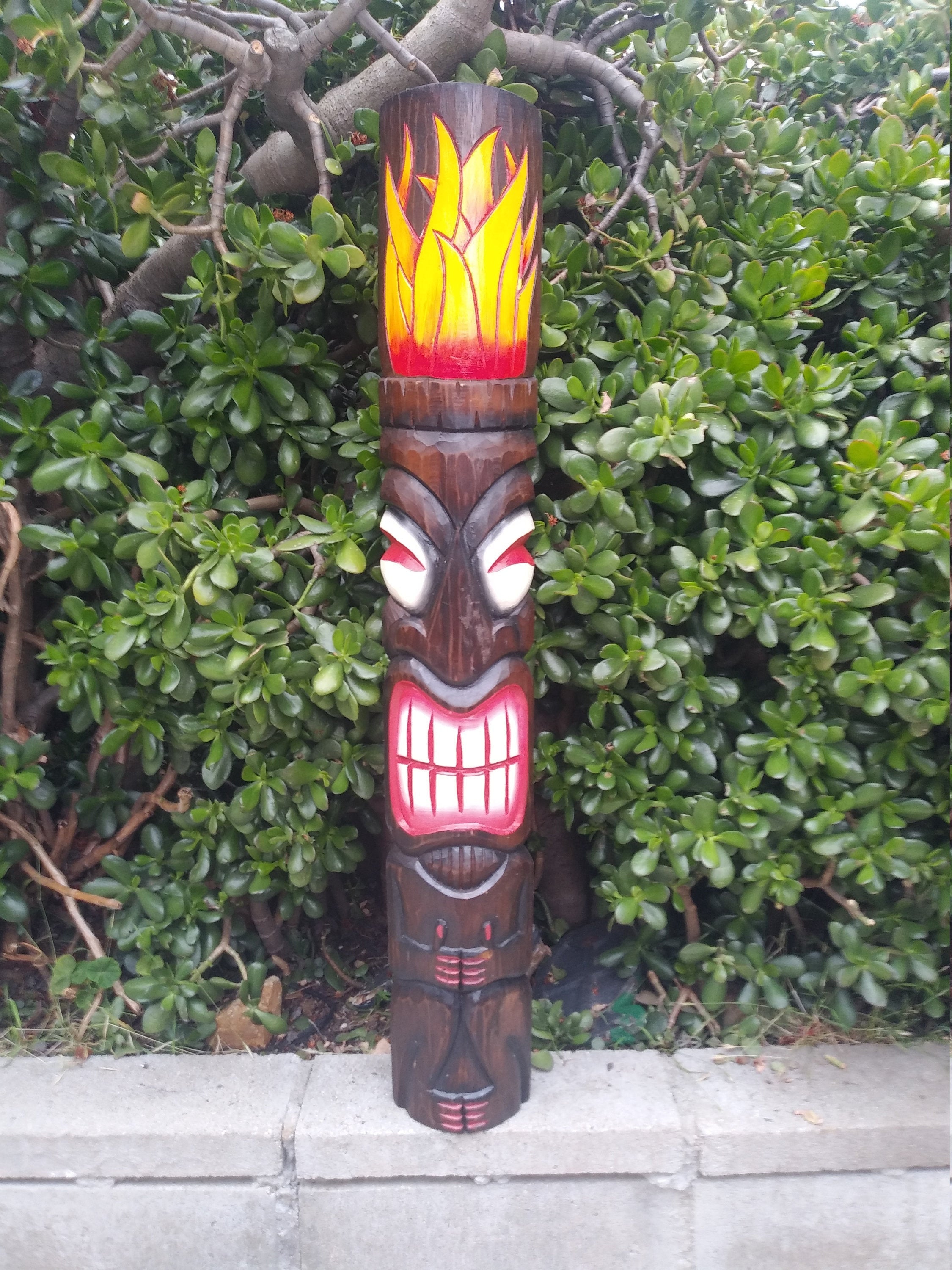 wooden tiki totem with flame-painted top and grinning carved face beside green shrubs