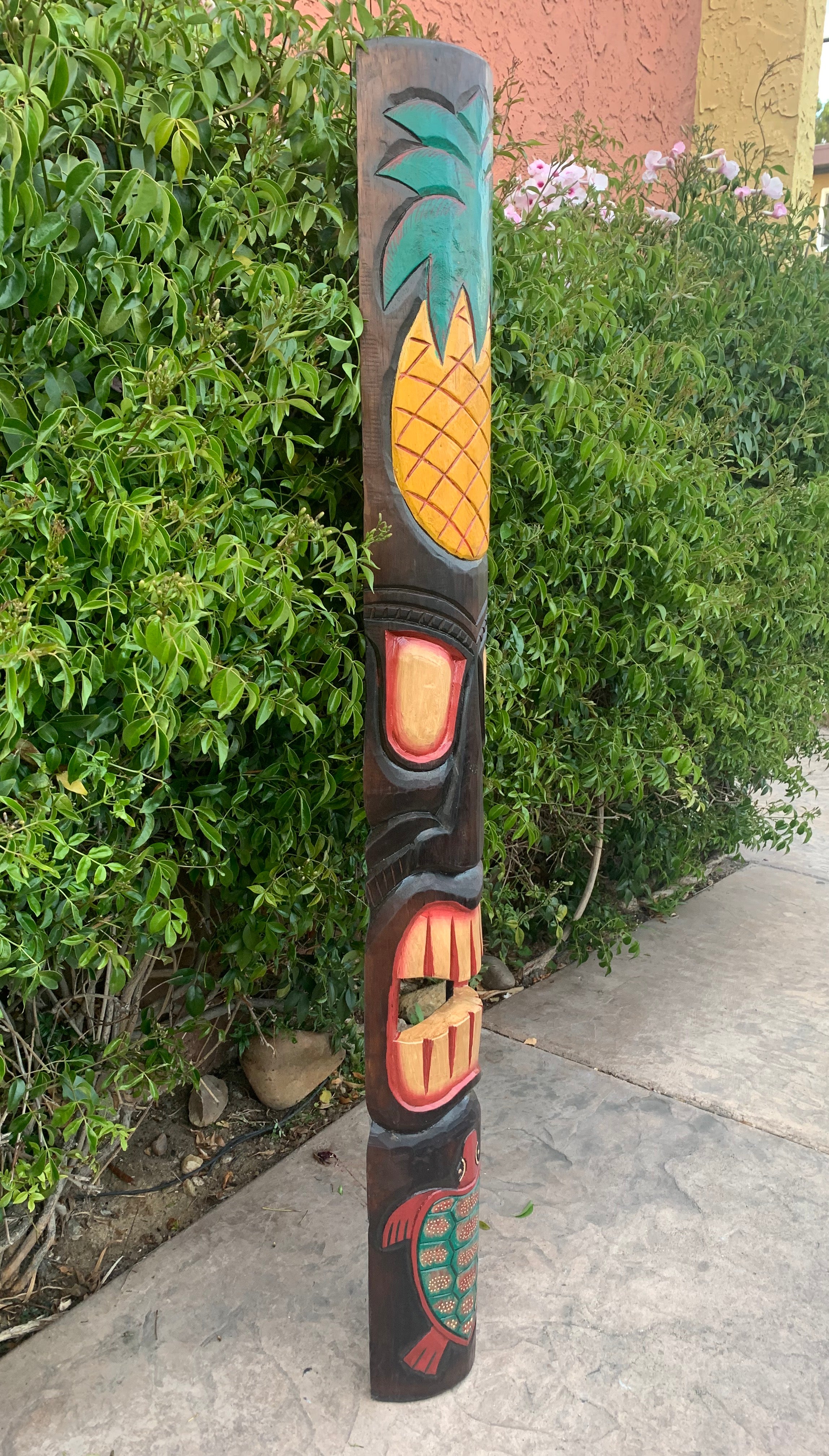 Carved wooden tiki totem pole featuring a pineapple at the top and a turtle design at the bottom beside a green hedge