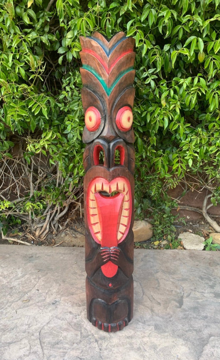 Wooden tiki totem statue with a large red mouth and round yellow eyes, set against green foliage.