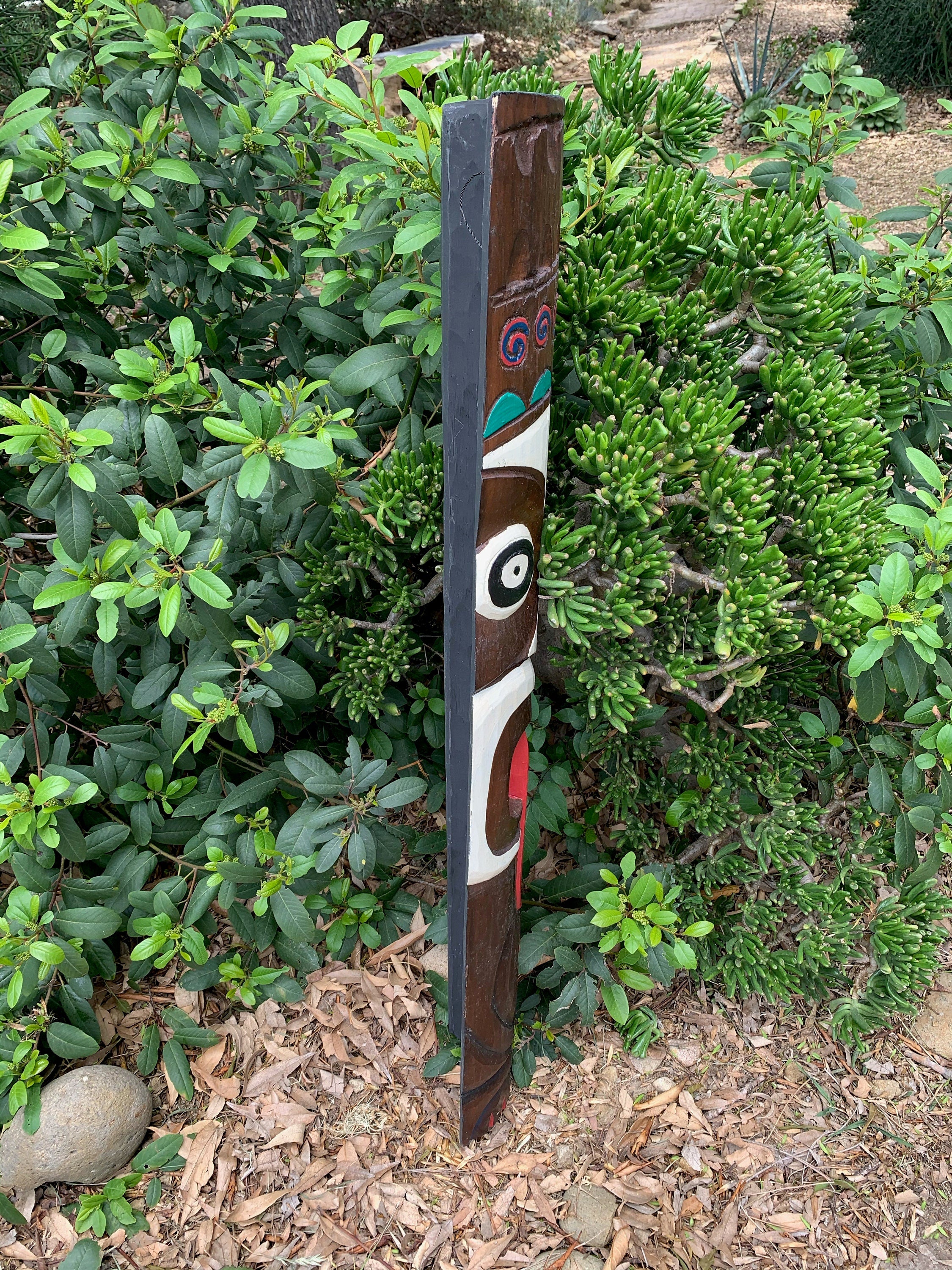 Vertical wooden totem pole carving with painted face in a garden among green shrubs.